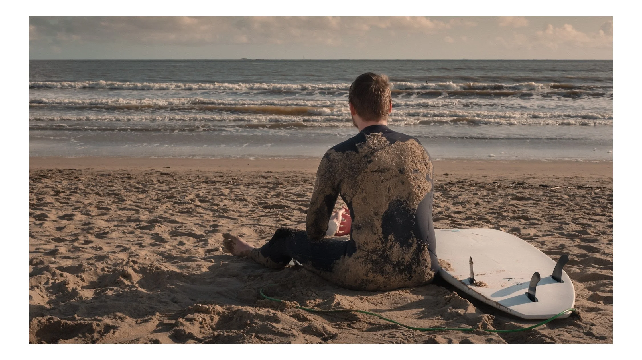 Un homme assis sur la plage, dos à la caméra, face à la mer, avec une planche de surf à côté, en fin d'après-midi.