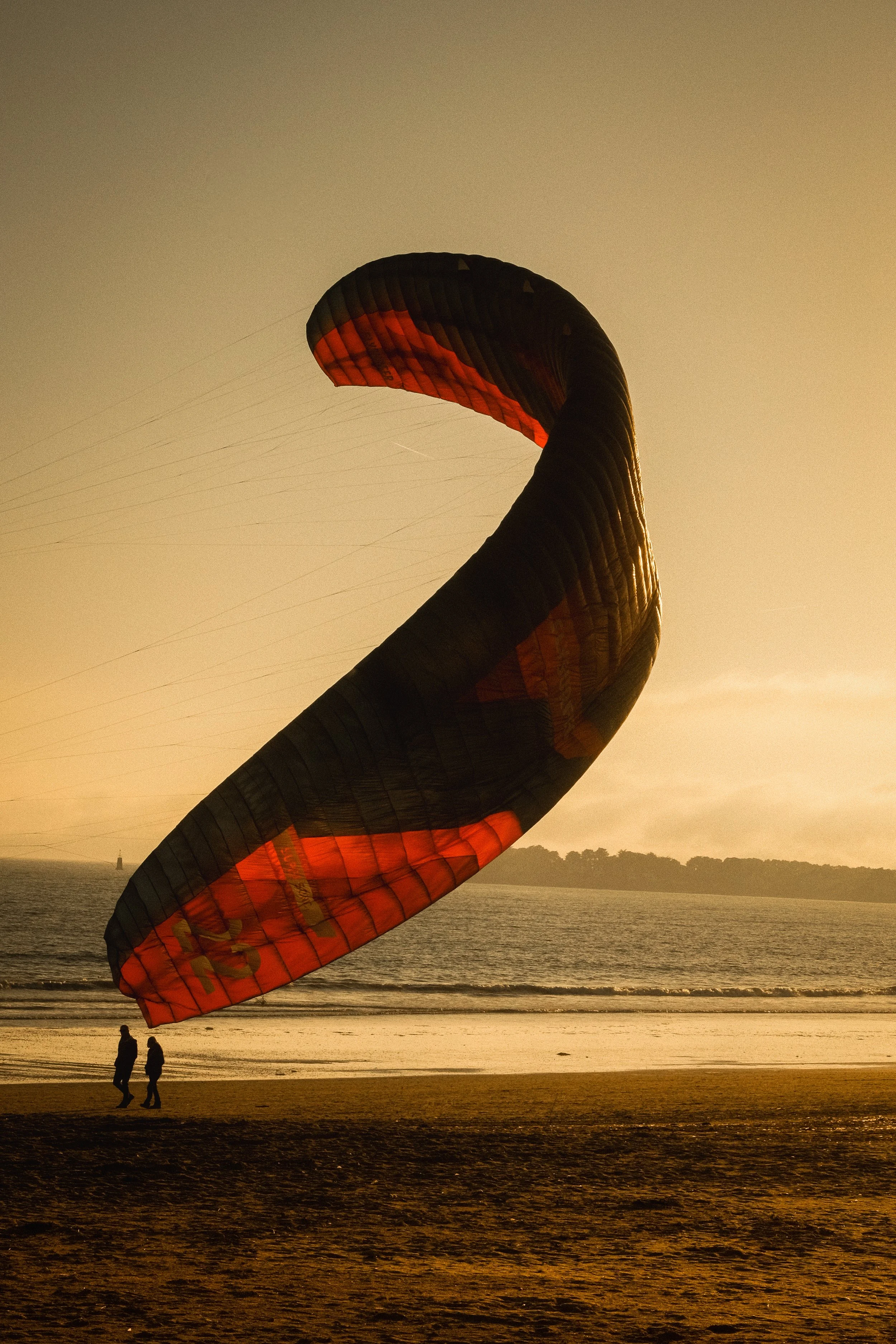 Deux personnes marchent sur la plage au coucher du soleil, avec une grande voile de parapente enroulée dans le sable, face à l'océan.