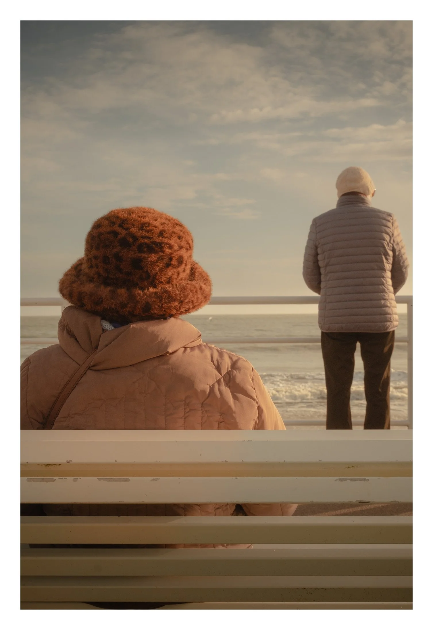 Deux personnes regardent l'océan depuis une jetée ou un quai, habillées pour le froid, avec un ciel nuageux.