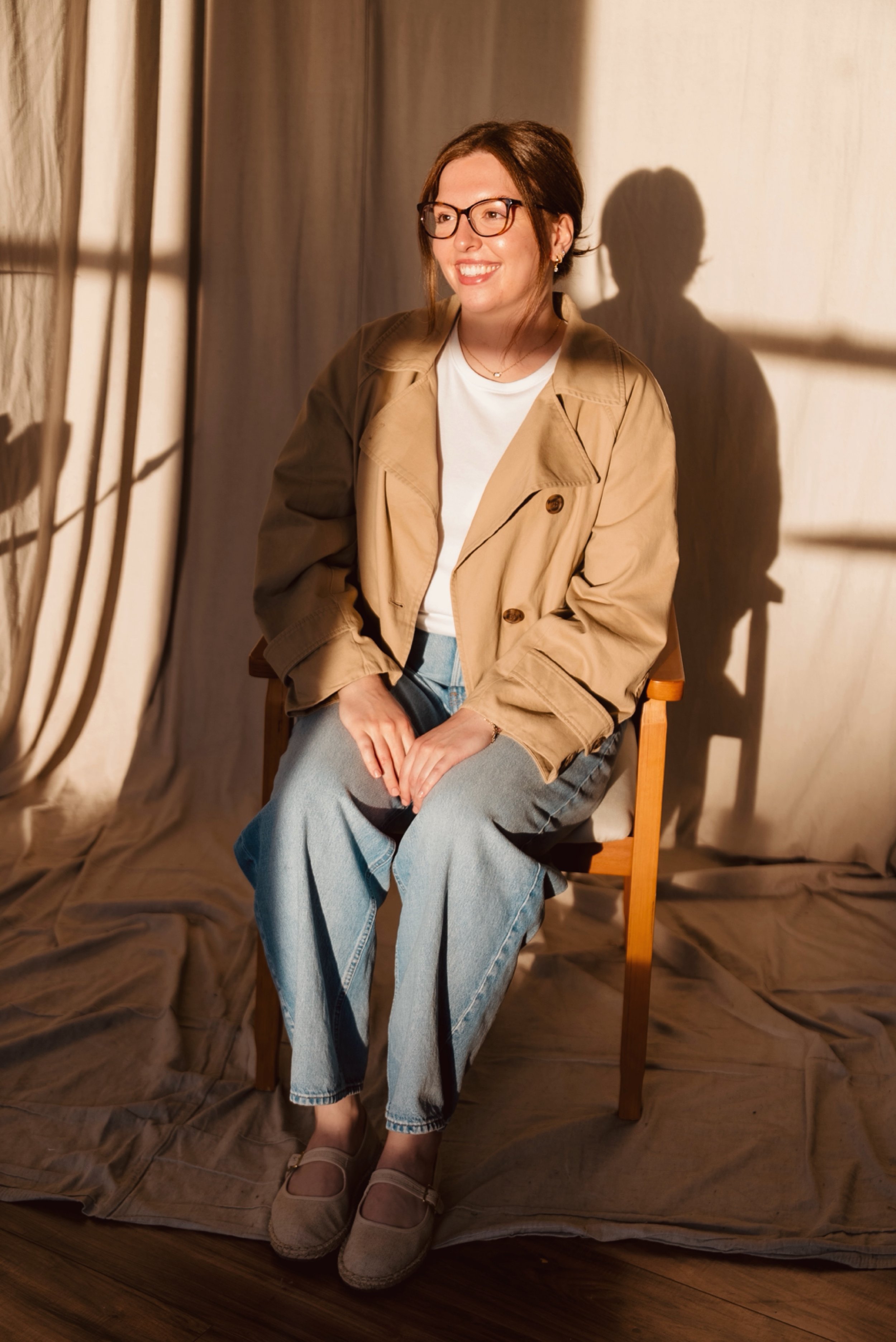 Smiling woman wearing glasses and a tan jacket seated in warm sunlight beside a curtained window