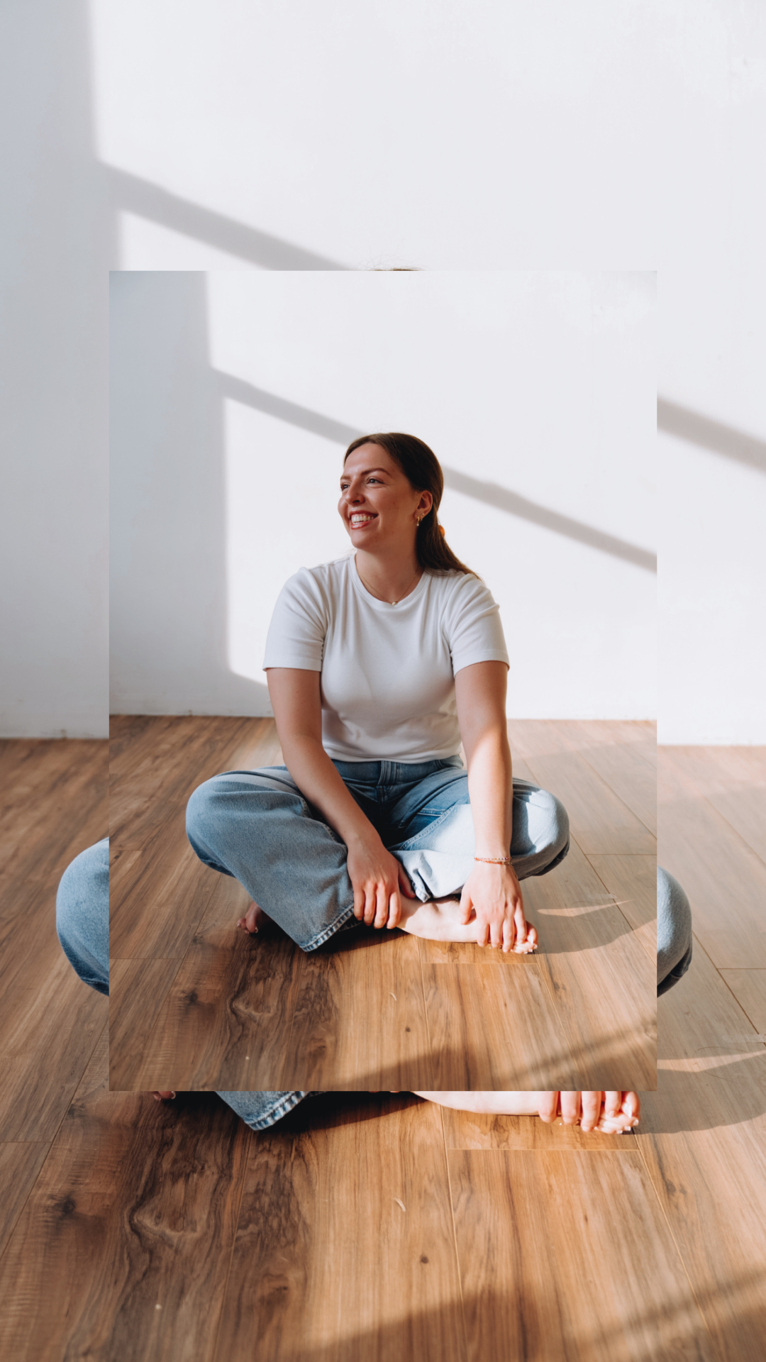 Smiling woman sitting cross-legged on a wooden floor in a sunlit room