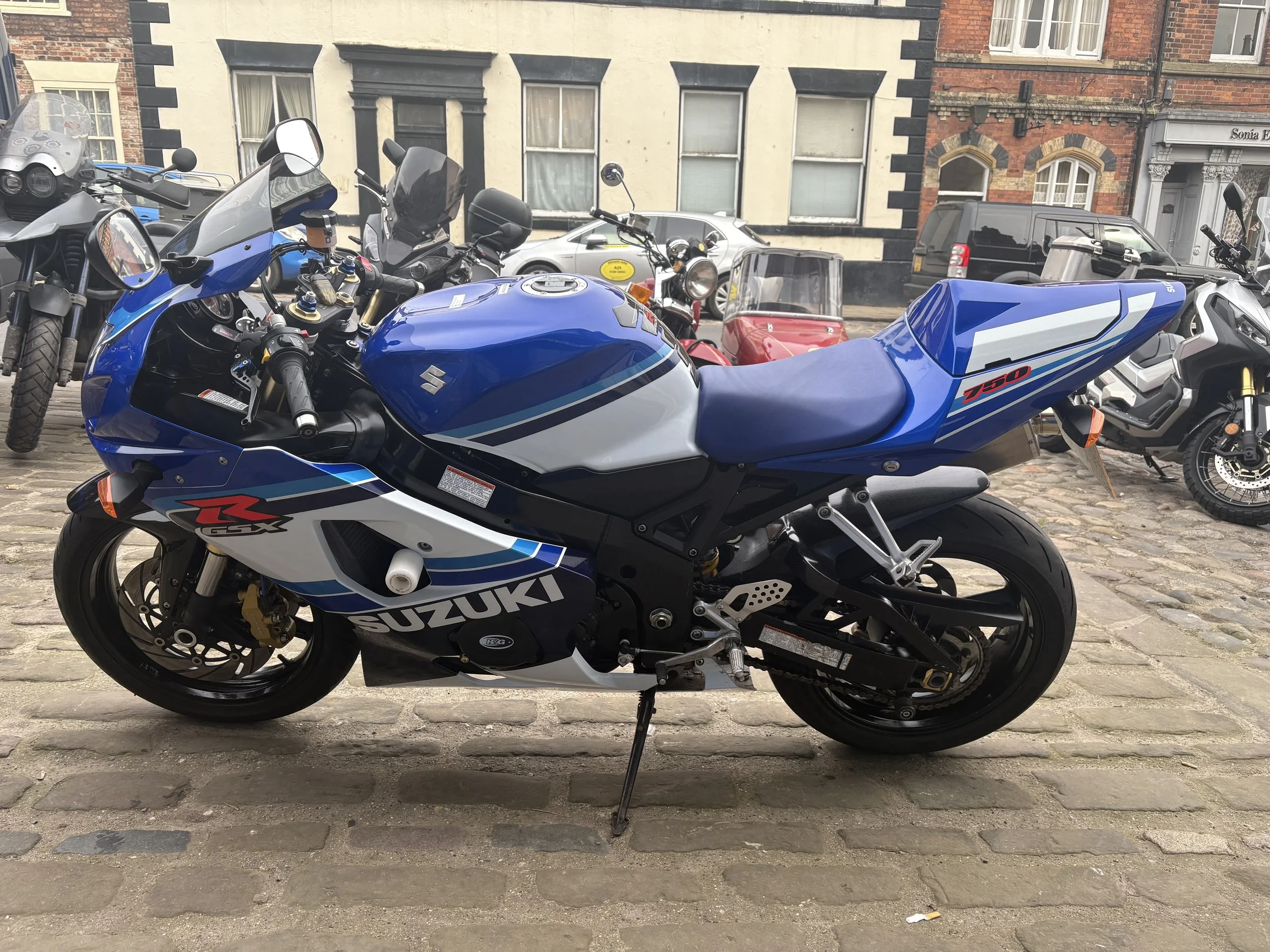 A blue and white Suzuki GSX-R 750 motorcycle parked on a cobblestone street, with other motorcycles and cars in the background.