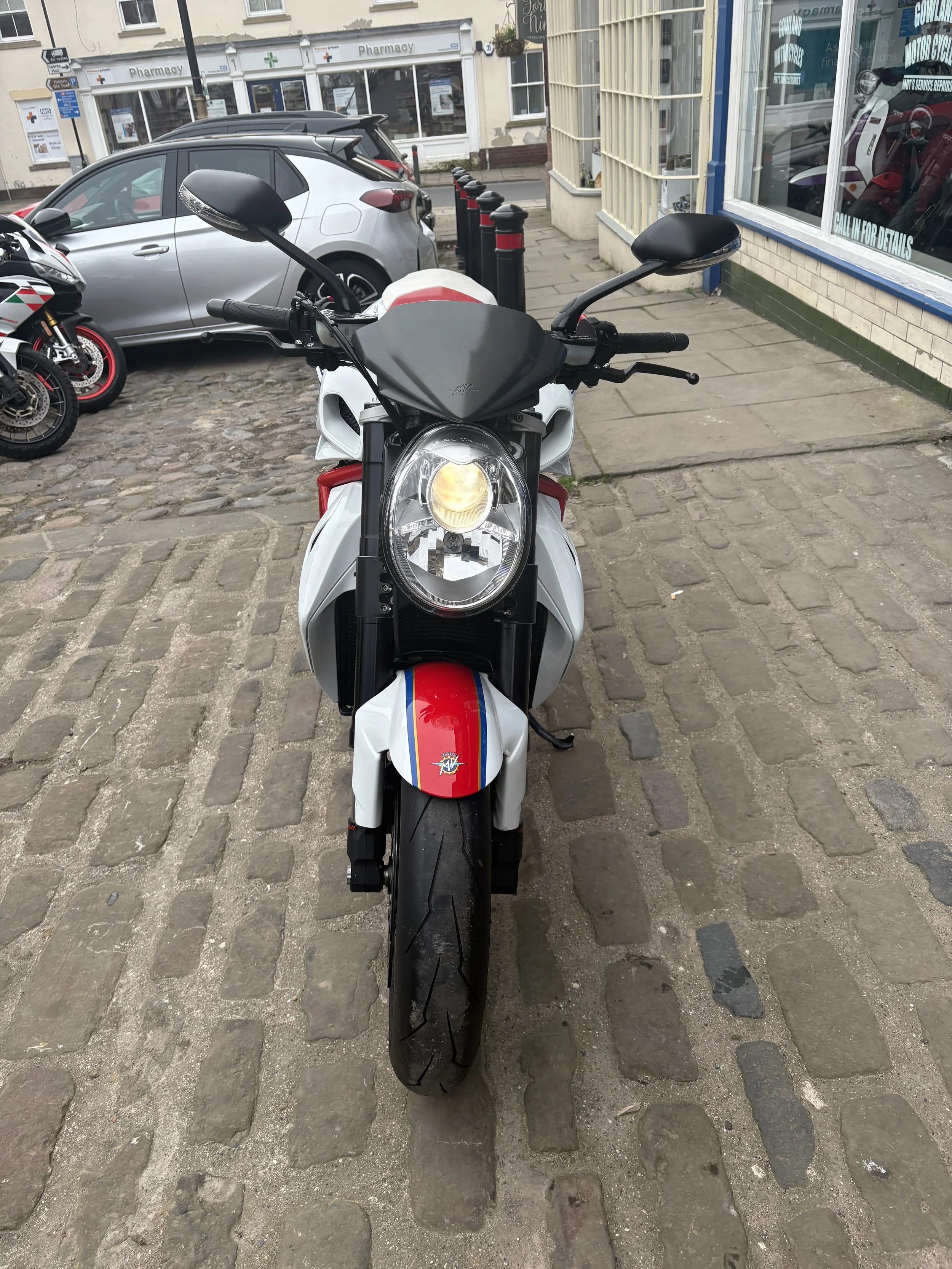 Front view of a white motorcycle with red, white, and blue stripes parked on a cobblestone sidewalk in front of a shop and other vehicles.