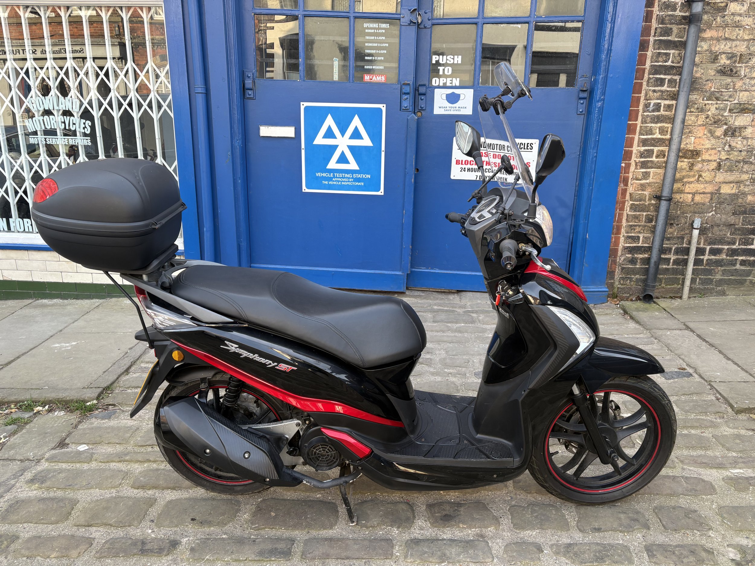 Black and red scooter parked on cobblestone sidewalk in front of a blue vehicle testing station door with signs and a small window.