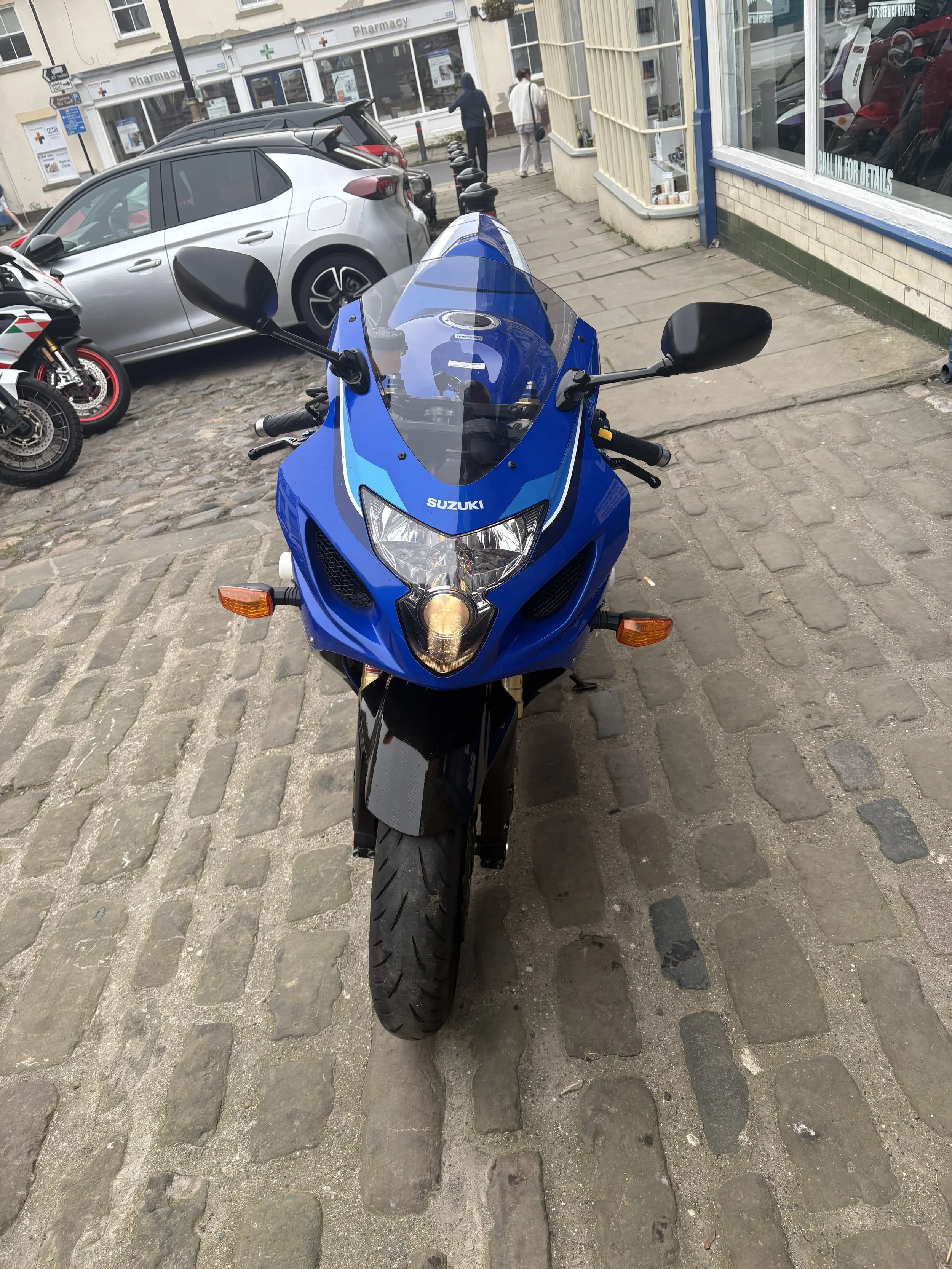 Front view of a blue Suzuki sport motorcycle parked on cobblestone sidewalk, with a pharmacy and other parked vehicles in the background.