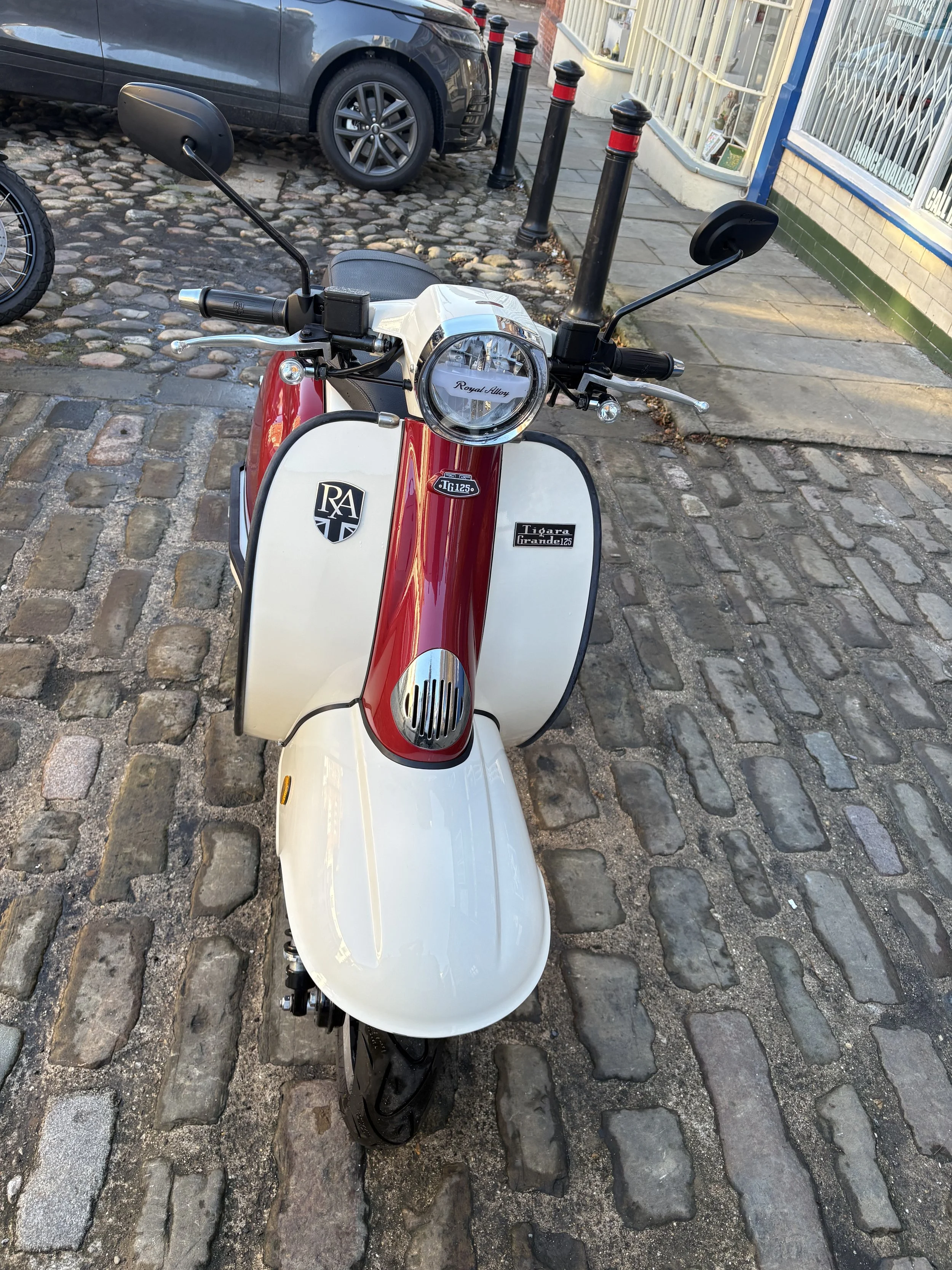 A vintage-style electric scooter parked on a cobblestone street, facing forward with a red and white color scheme, black handlebars, and a round headlight.