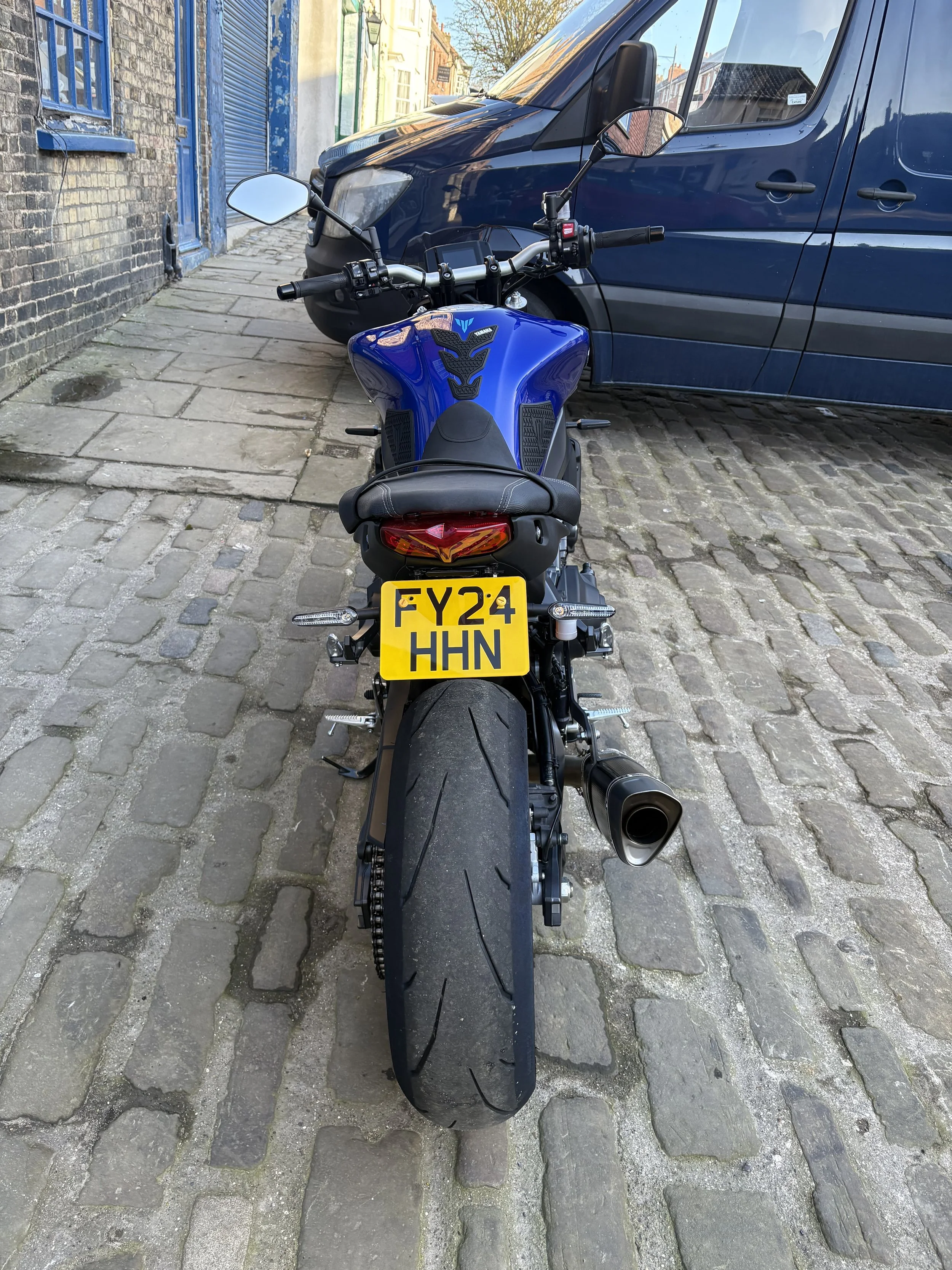 Blue motorcycle parked on a cobblestone street with a dark blue van behind it. The motorcycle has a yellow license plate with the number FY24 HHN and a single exhaust pipe on the right side.