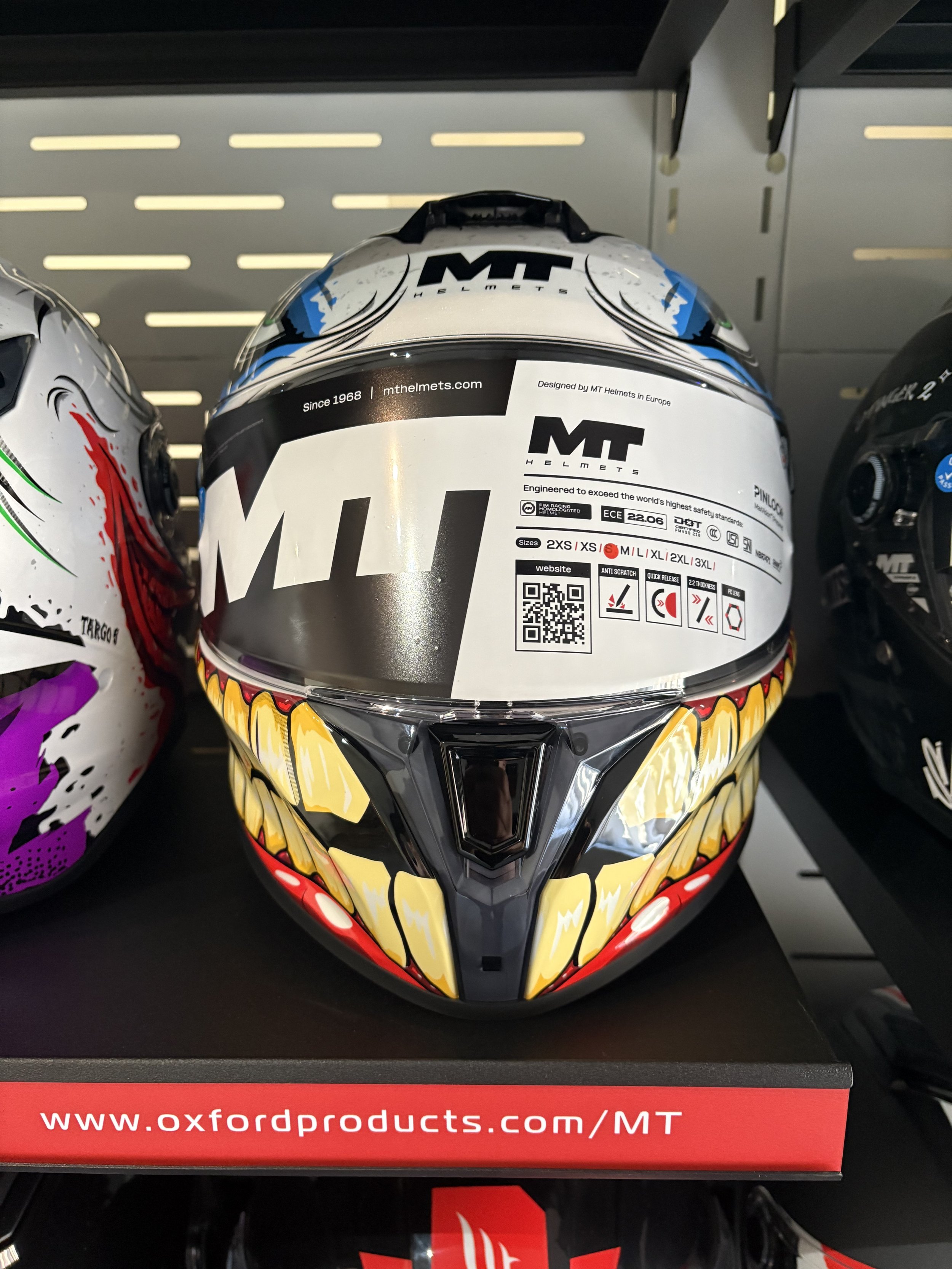 An MT motorcycle helmet with a fierce design resembling an open mouth with sharp teeth, primarily in color yellow with black and red accents. The helmet is displayed on a shelf, next to other helmets.