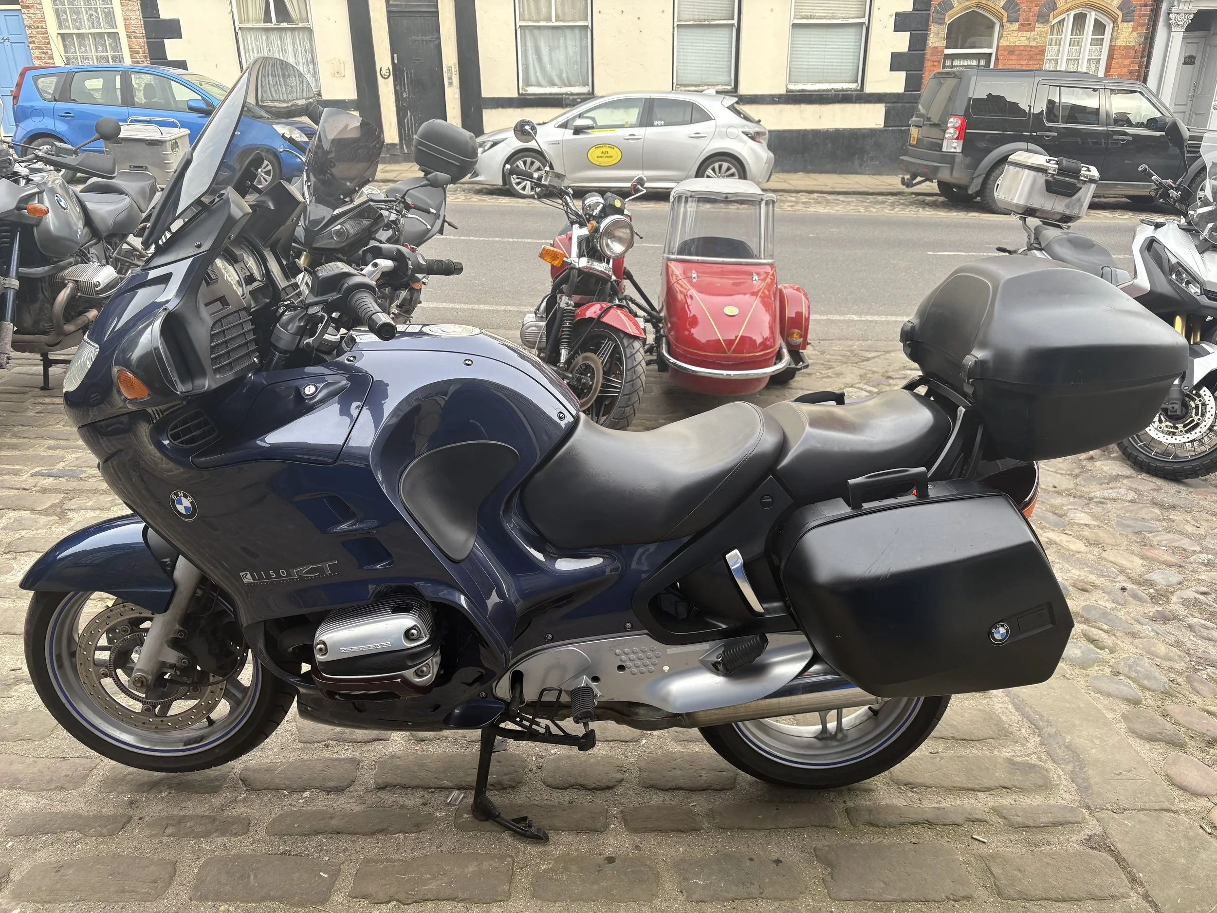 A blue BMW motorcycle with side panniers parked on cobblestone street, with other motorcycles, a red motorcycle with a sidecar, and cars visible in the background.