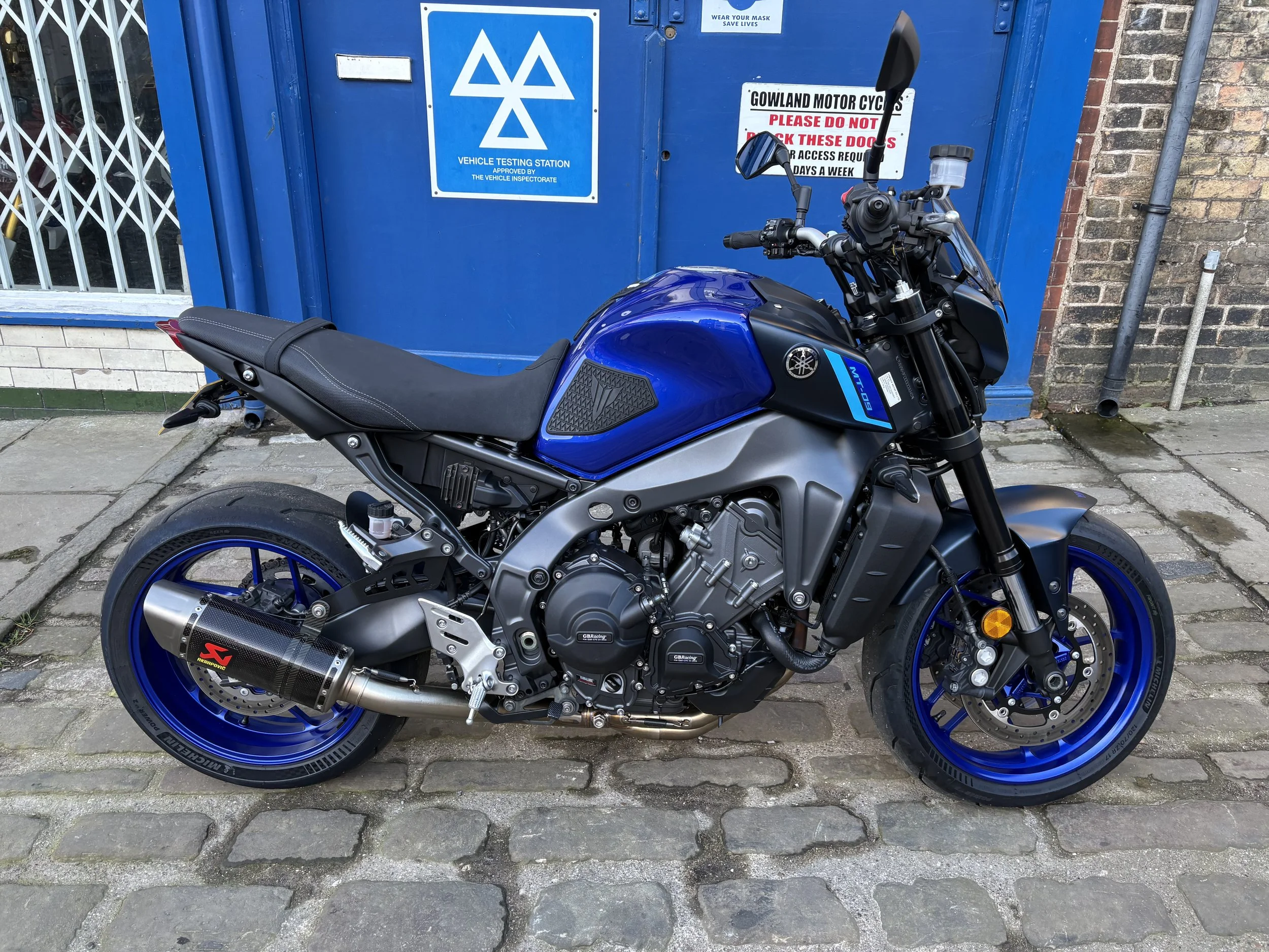 Blue Yamaha motorcycle parked on brick-paved sidewalk in front of a blue vehicle testing station. The motorcycle has a black seat, black handlebar grips, blue wheels, and a black exhaust.