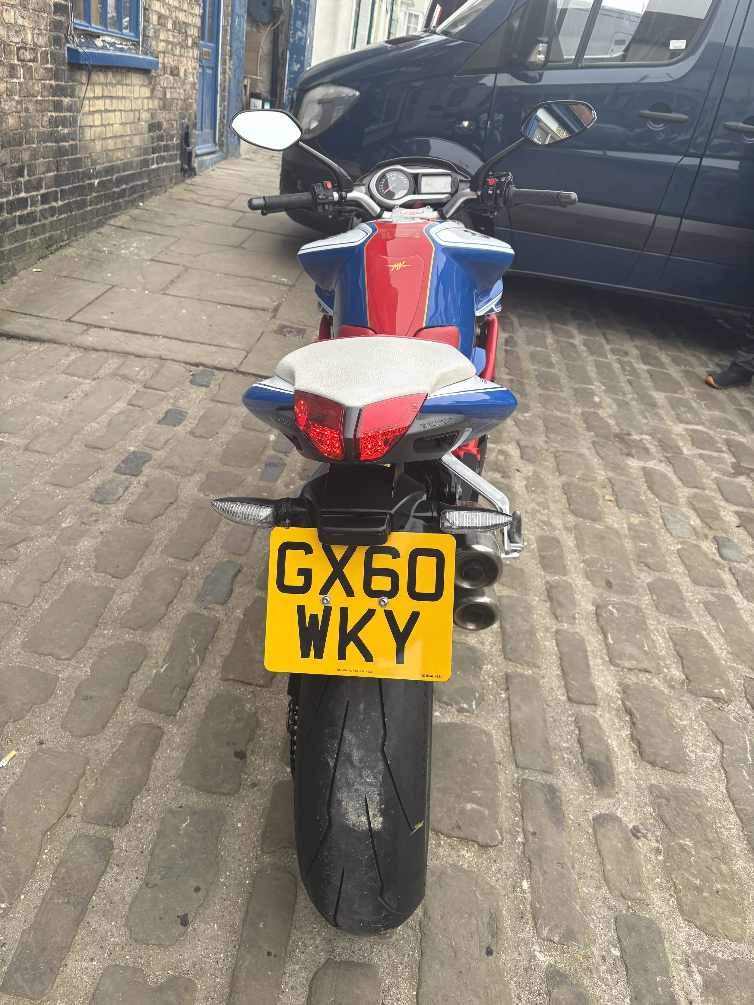 Back view of a motorcycle with a yellow license plate reading 'GX60 WKY', parked on a brick-paved street next to a dark blue delivery van.