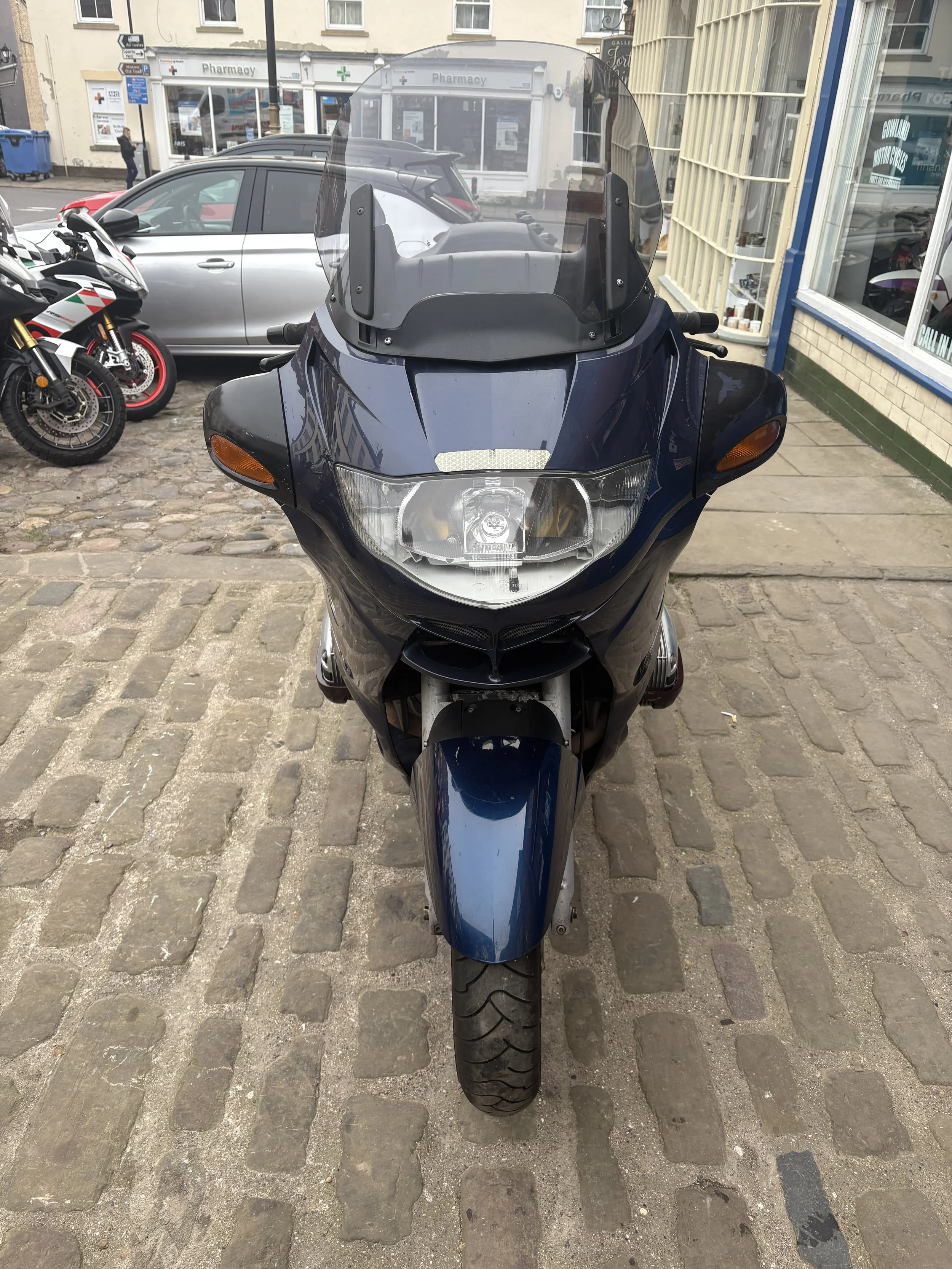 Front view of a dark blue motorcycle parked on a cobblestone street, with a storefront, other vehicles, and motorcycles in the background.