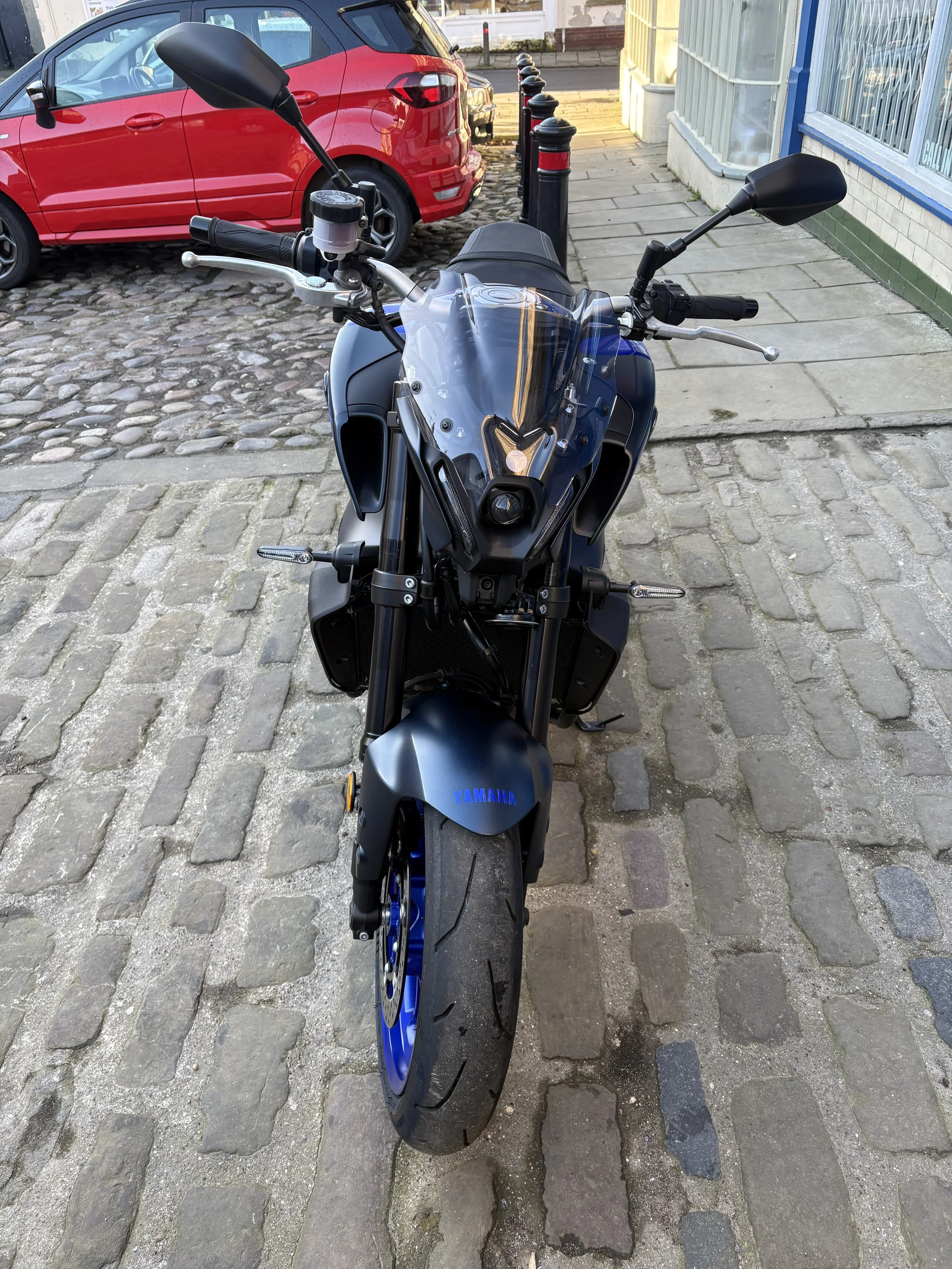 Front view of a black Yamaha motorcycle parked on a cobblestone street, with a red car and shop storefront in the background.