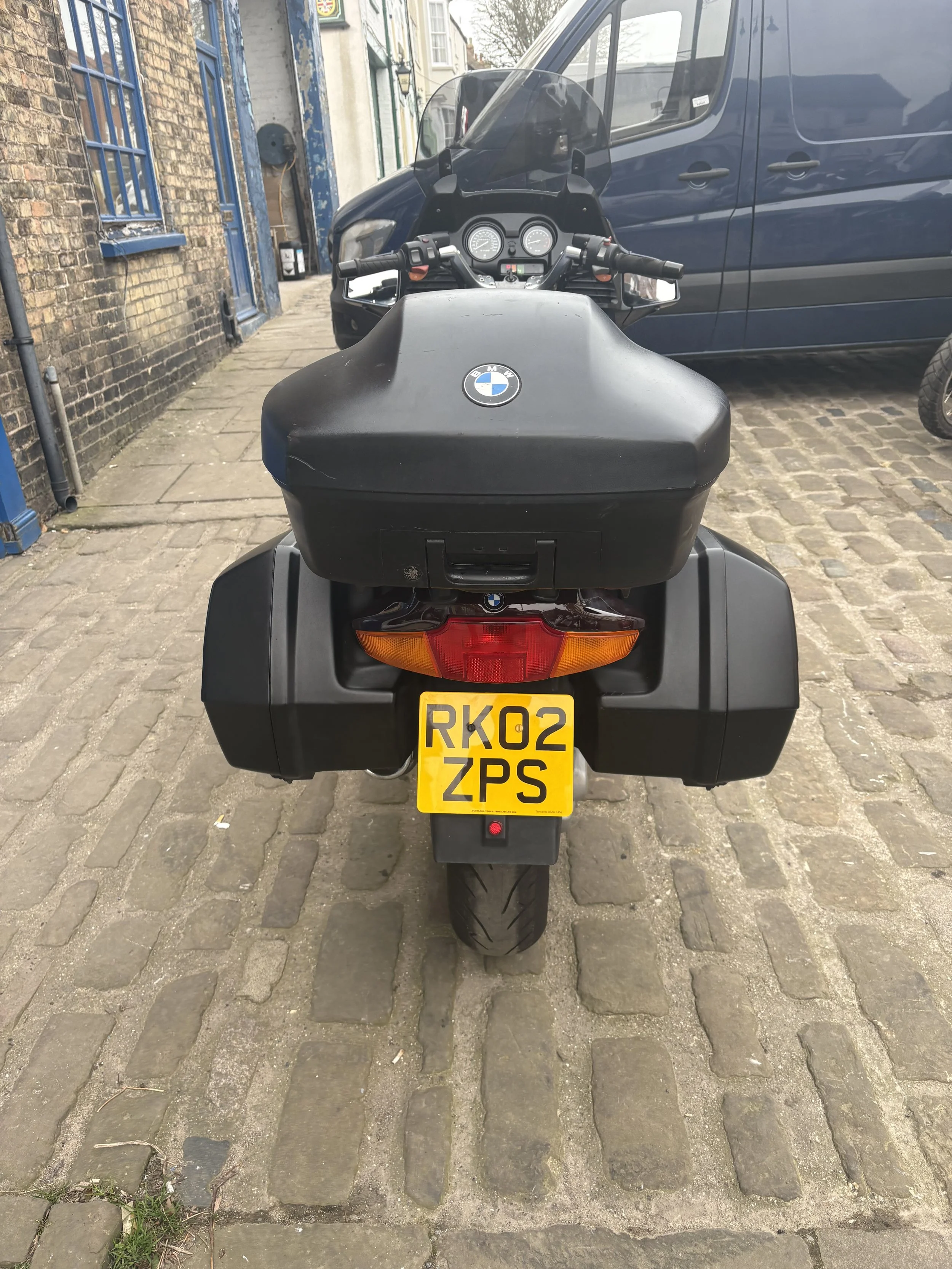 Rear view of a black BMW motorcycle with a yellow UK license plate on a cobblestone street, parked next to a brick building and a blue van.