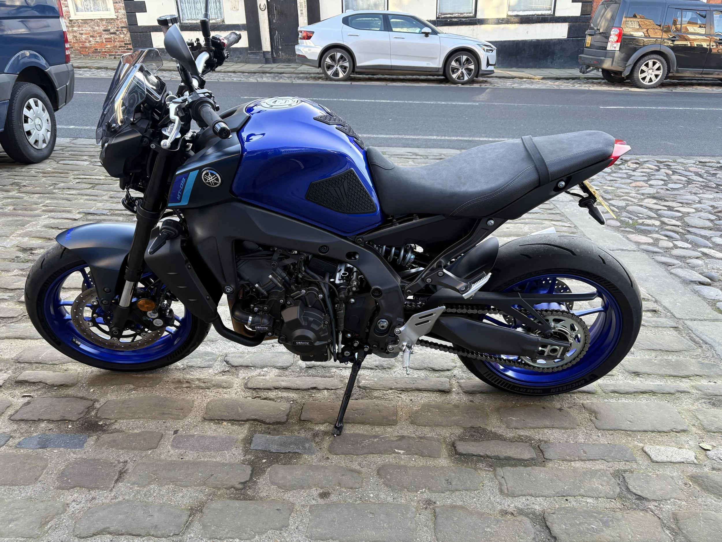 Blue Yamaha motorcycle parked on cobblestone street with cars and brick buildings in the background.