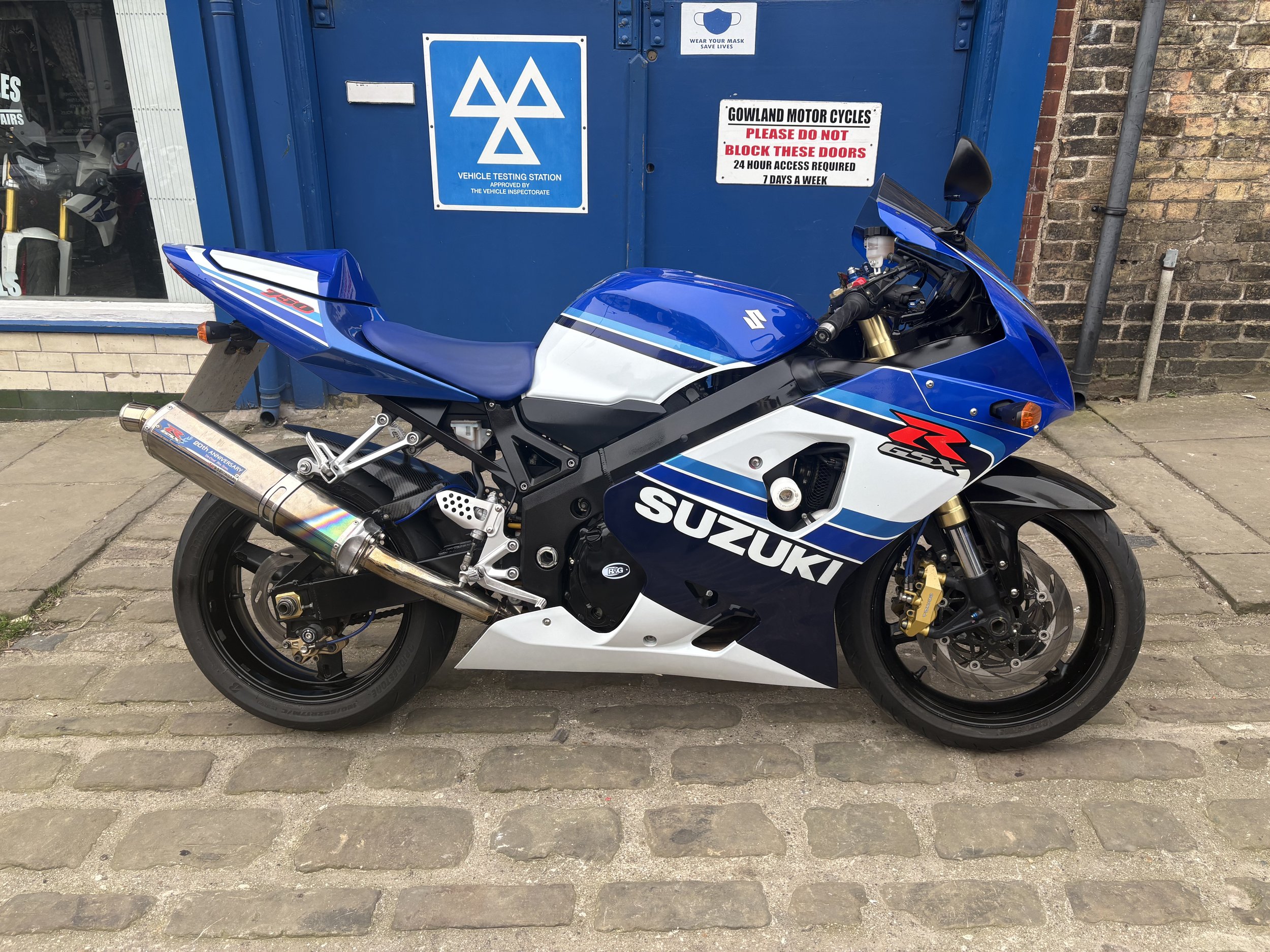 Blue and white Suzuki GSX-R motorcycle parked on a cobblestone sidewalk in front of a blue vehicle testing station door with signs.
