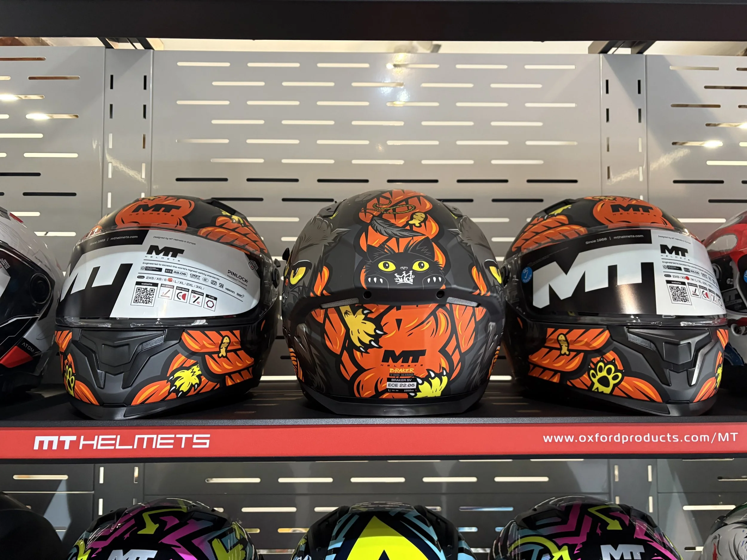 Display of three motorcycle helmets on a shelf featuring black, orange, and yellow Halloween-themed designs, with two helmets on the sides having red and black badging and the middle helmet with a black cat and pumpkins.
