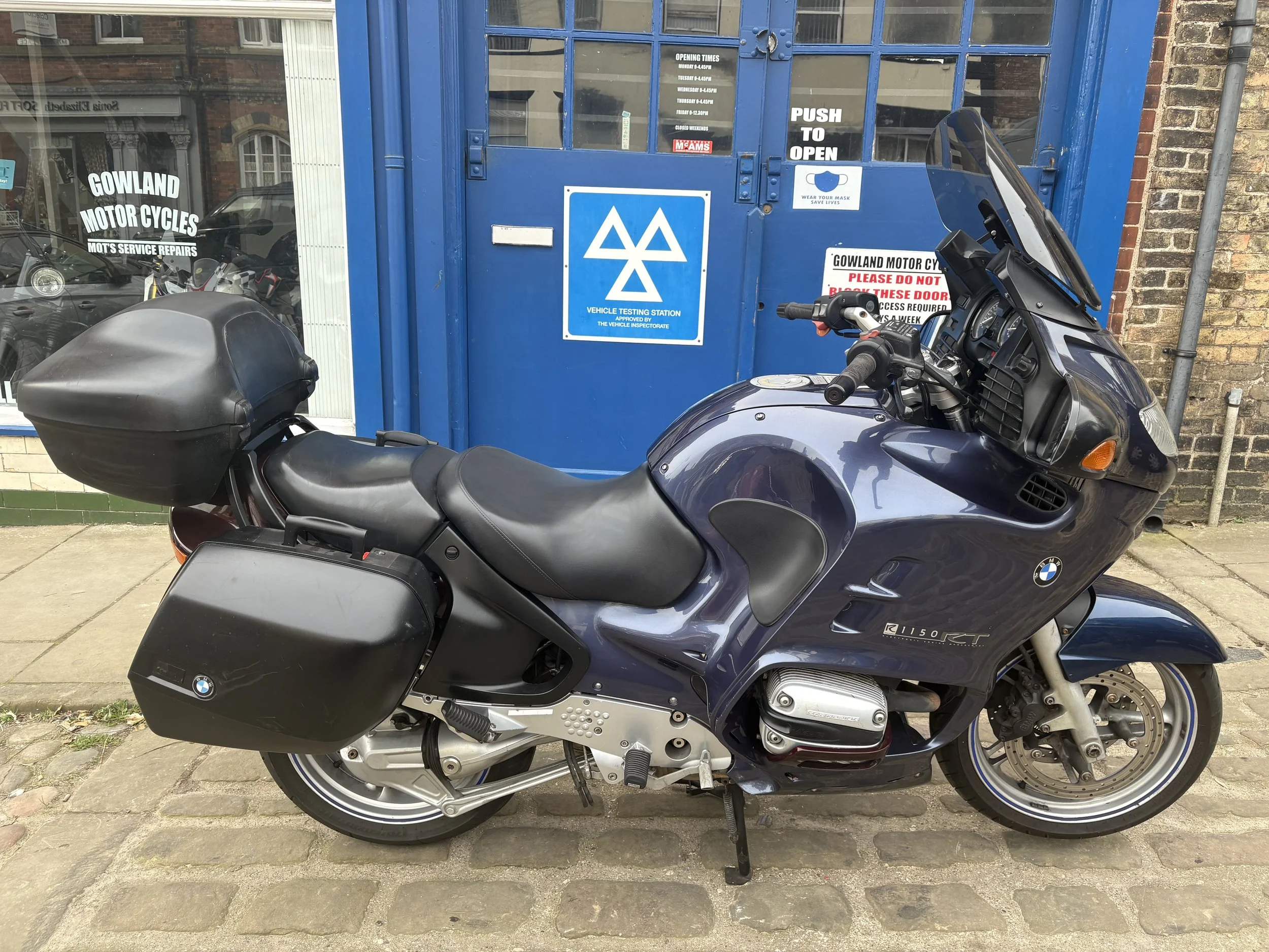 A blue BMW motorcycle parked outside a vehicle testing station with a black top case and side panniers. The station has a blue door and signs, including a MOT service repair shop and a vehicle testing station logo.