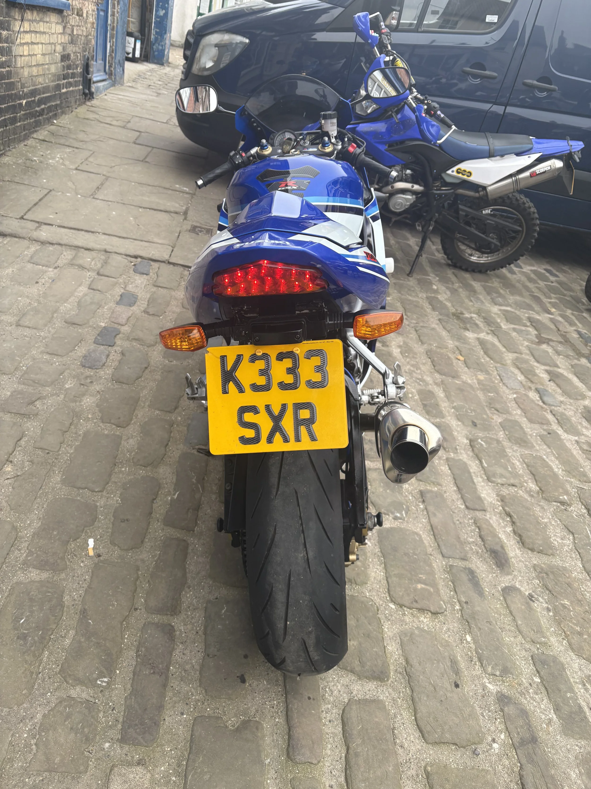 Rear view of a blue motorcycle with a yellow license plate reading 'K333 SXR', parked on a cobblestone street, with a black car and a dirt bike in the background.
