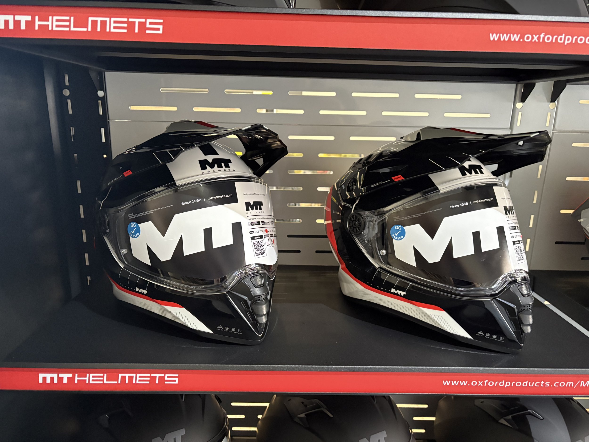 Two black and white motorcycle MT helmets with tinted visors on a retail shelf, branded with 'MT Helmets' logos.