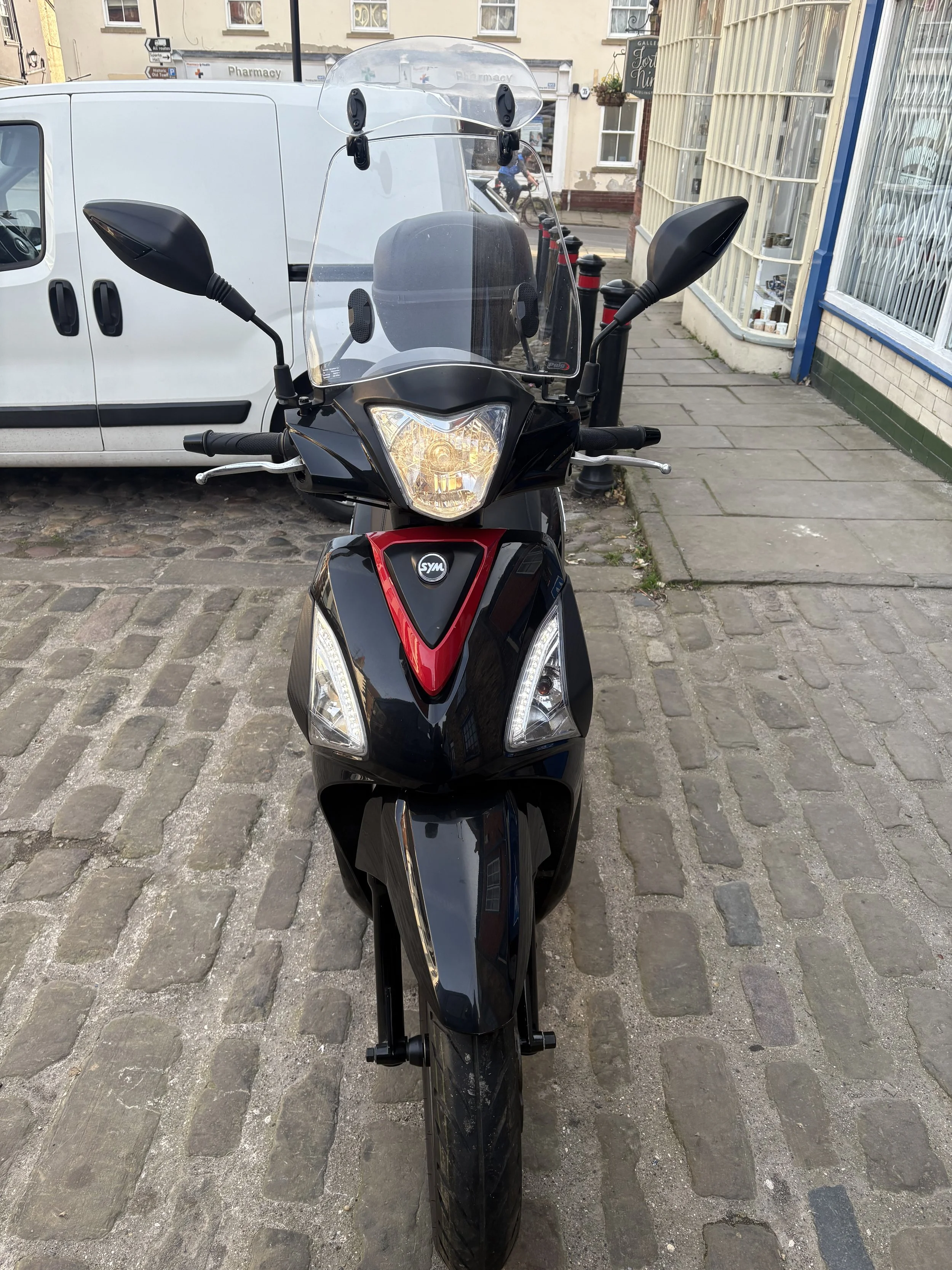 Black scooter with a clear windshield parked on a cobblestone street in front of a white van and storefronts.