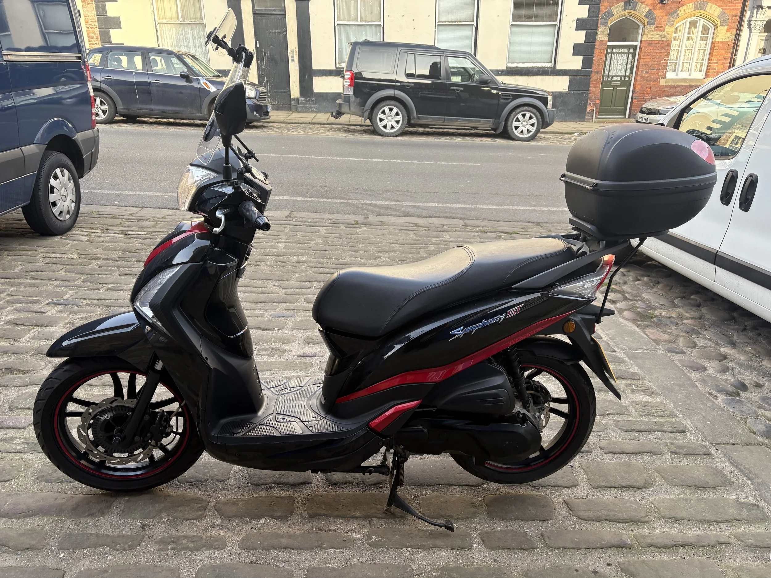 Black scooter with a top box parked on a cobblestone street in front of a row of cars and historical buildings.