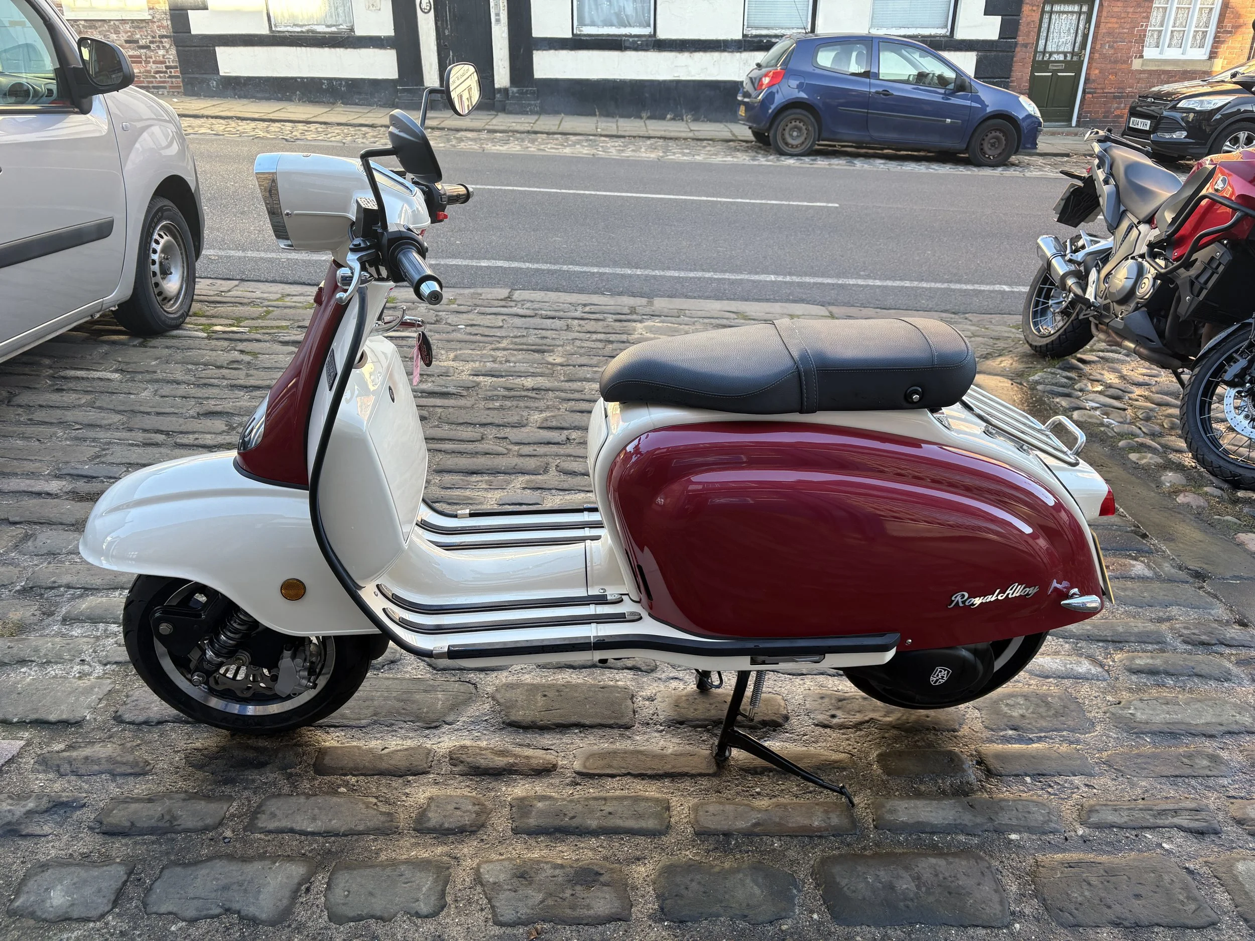 A red and white vintage-style Vespa scooter parked on cobblestone street, with a black seat, side stand, and a rear rack.