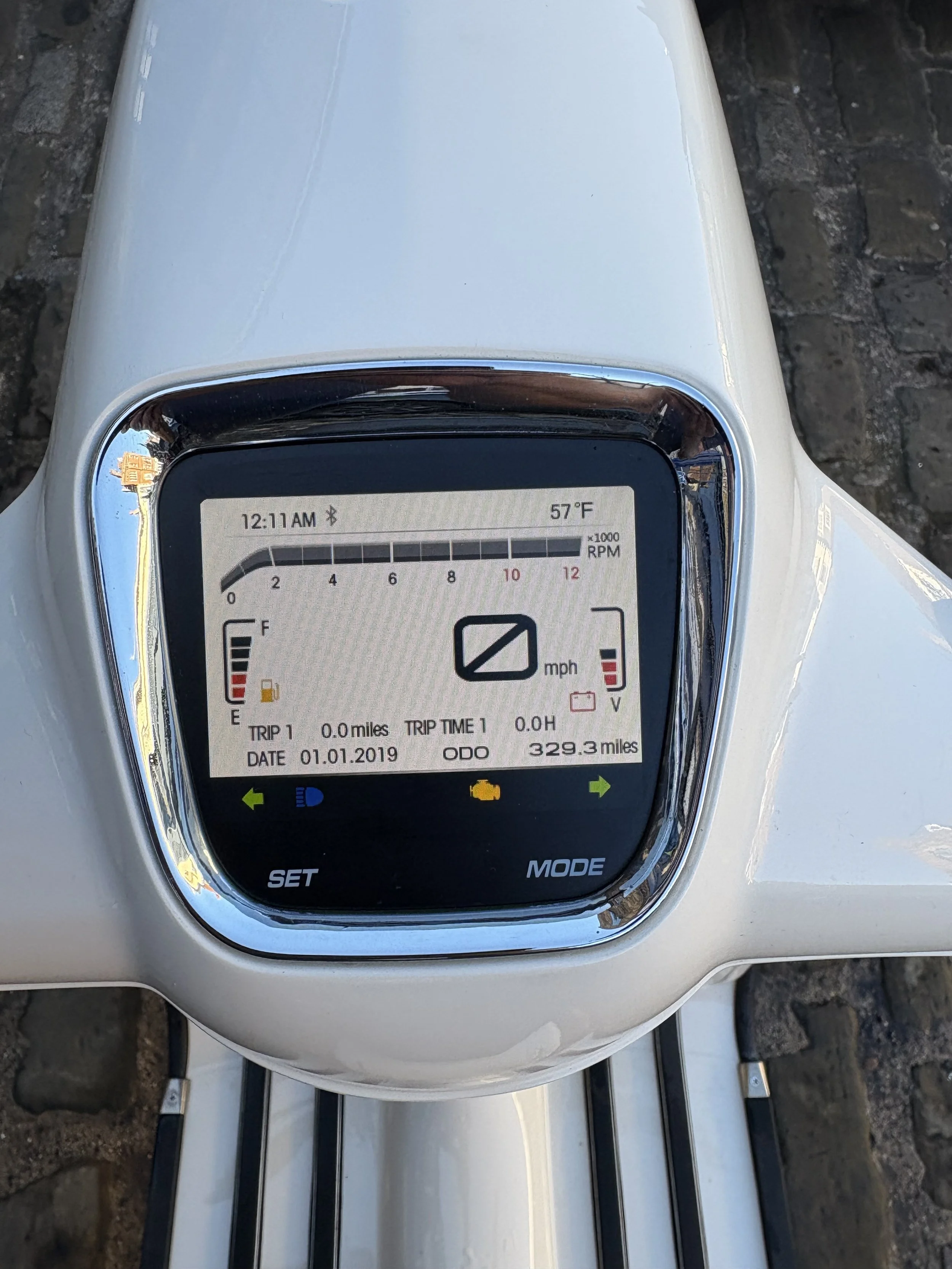 Digital dashboard display of a scooter showing 0 mph, trip distance of 0 miles, and total mileage of 329.3 miles. The screen shows the date as 01.01.2019, time as 12:11 AM, temperature as 57°F, and the battery and warning indicators.