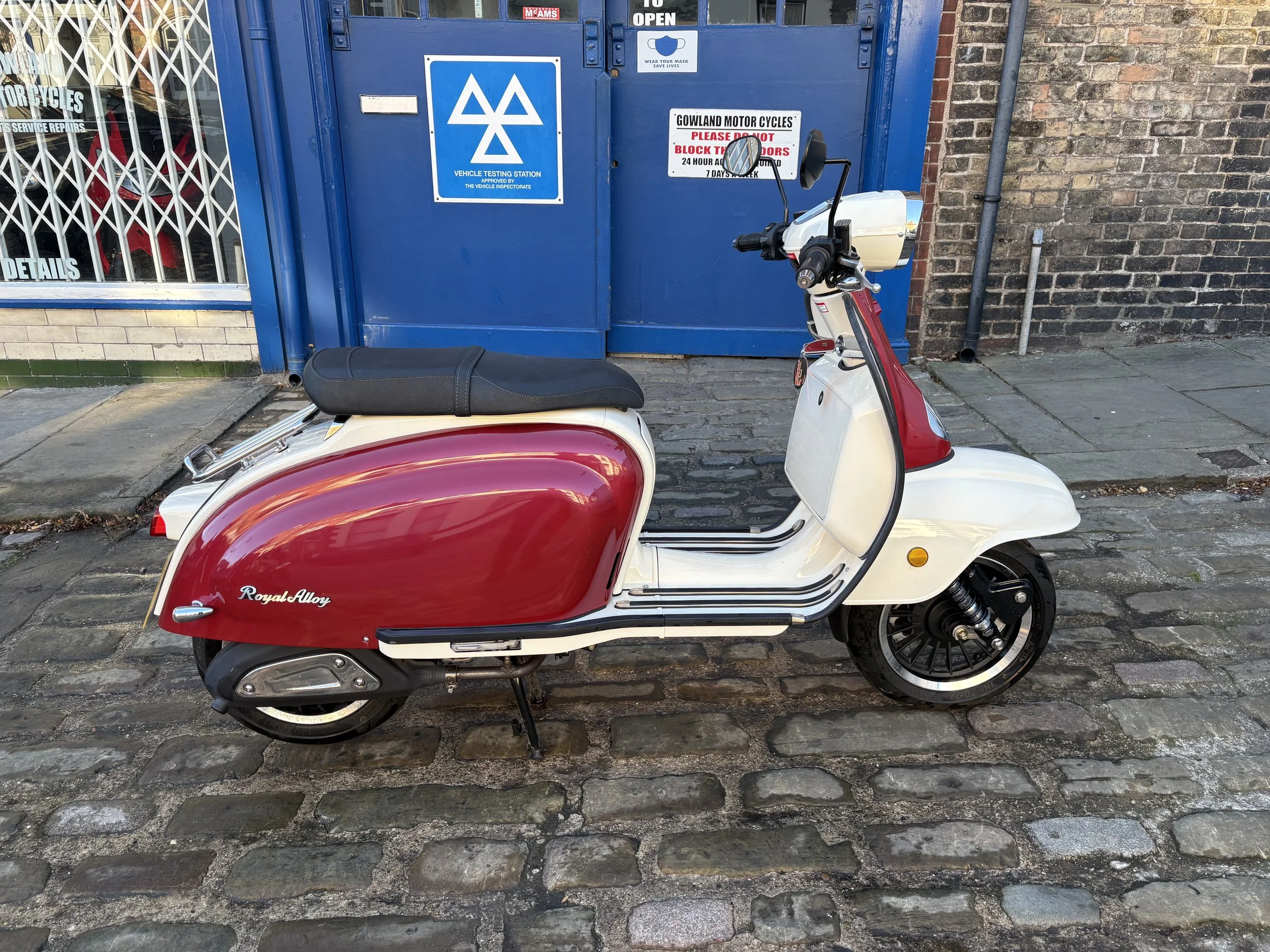 A vintage red and white Royal Alloy scooter parked on a cobblestone street in front of a blue vehicle testing station gate. The scooter has a black seat and black wheels, with a small mirror on the handlebar.