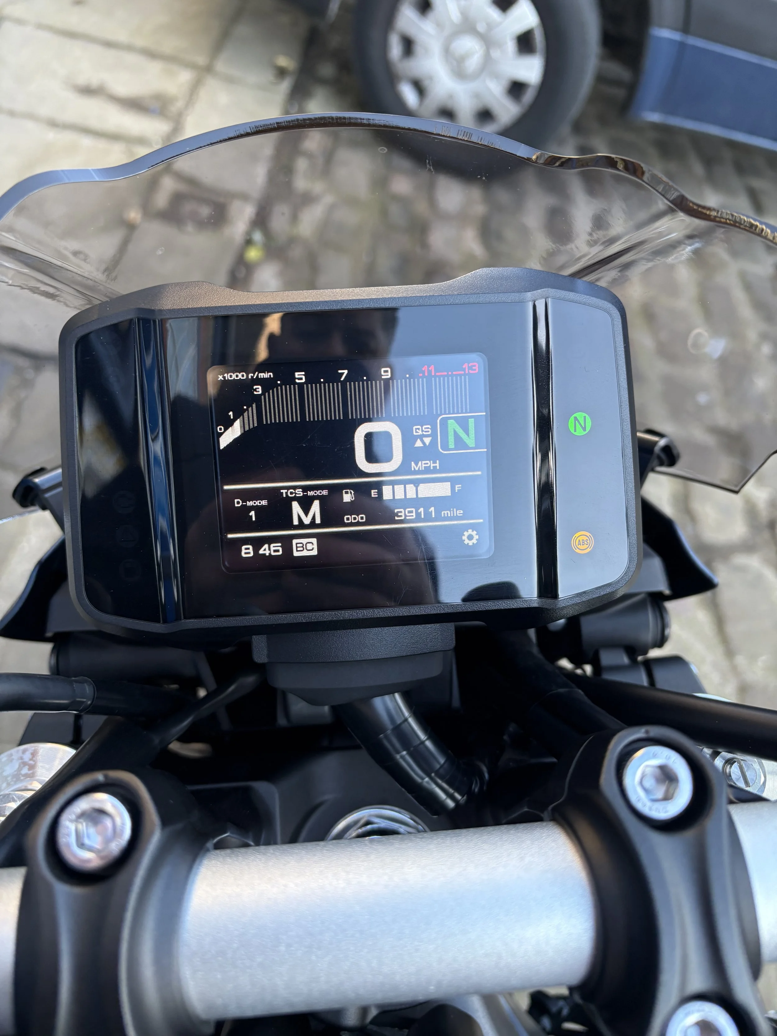 Close-up of a motorcycle's digital dashboard displaying a speed of 0 MPH, full fuel, 3911 miles odometer, with a clear windshield and a blurred background of a parked car and a paved driveway.