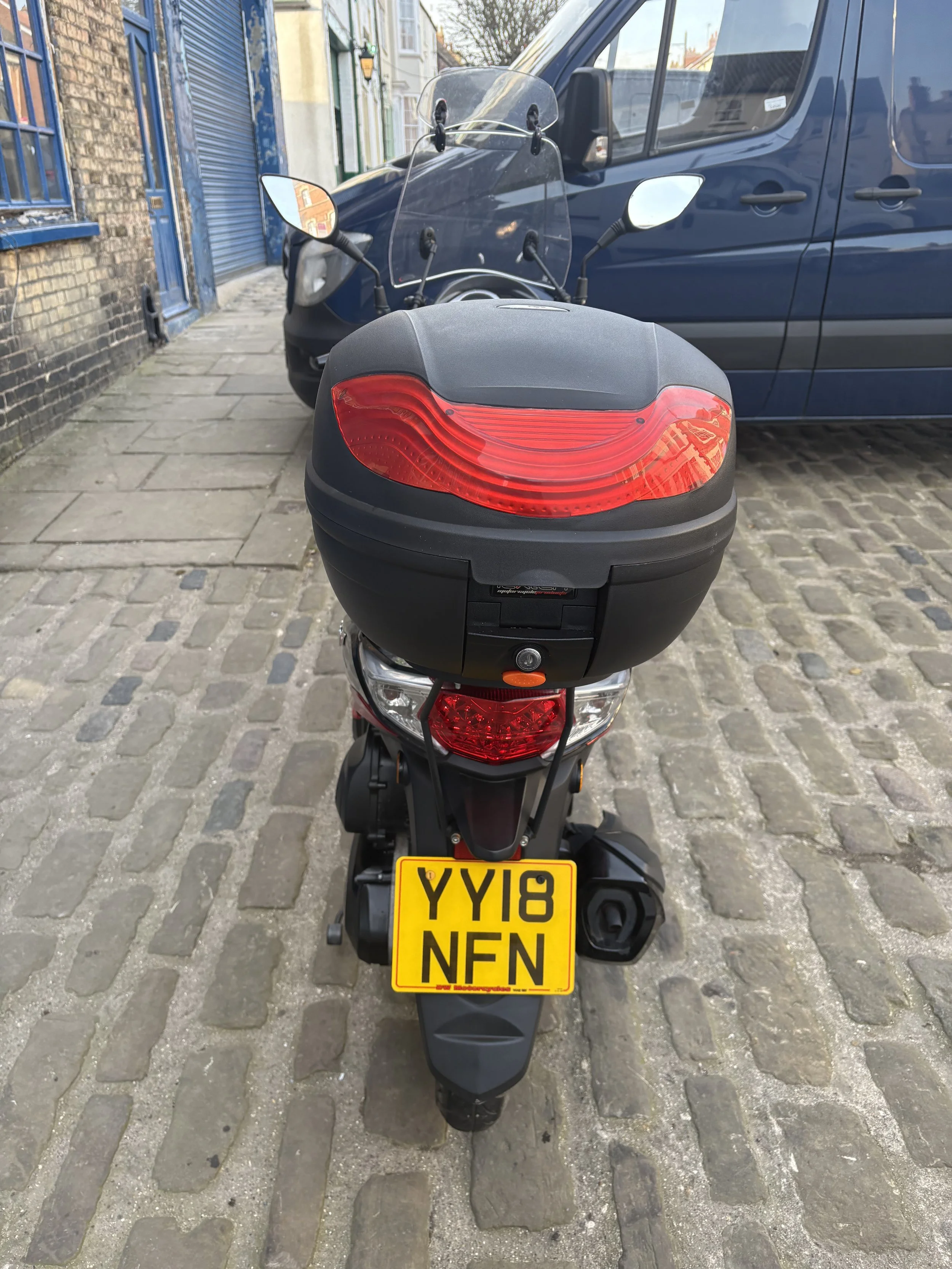 Rear view of a black scooter with a top case, parked on a cobblestone street next to a blue van and a brick building.