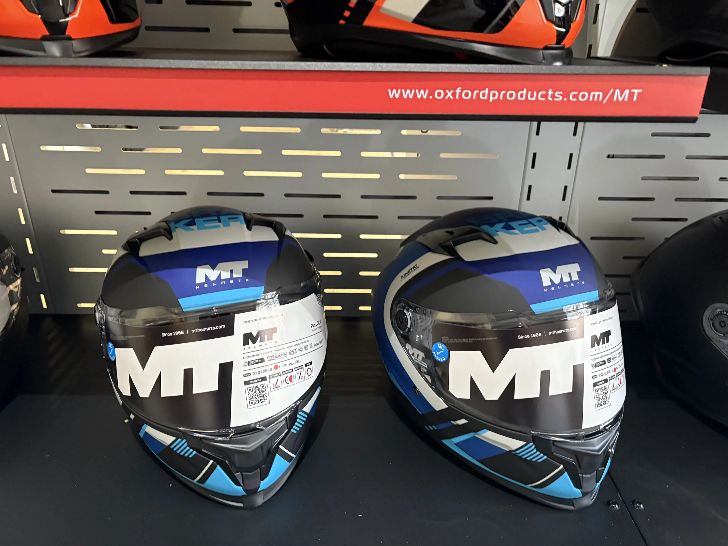 Two MT motorcycle helmets on display, black, blue, and white, with the brand 'MT' logo, placed on a black shelf in a store.