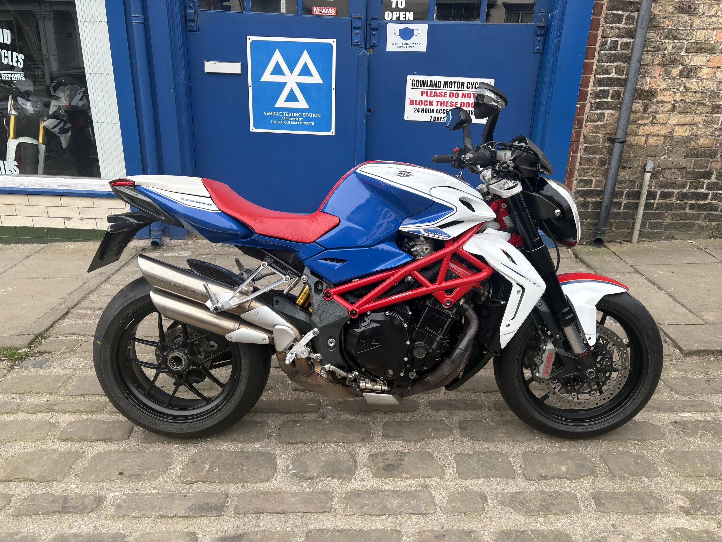 A blue, white, and red sport motorcycle parked on a cobblestone street in front of a blue vehicle testing station with signs and a window reflecting motorcycles inside.