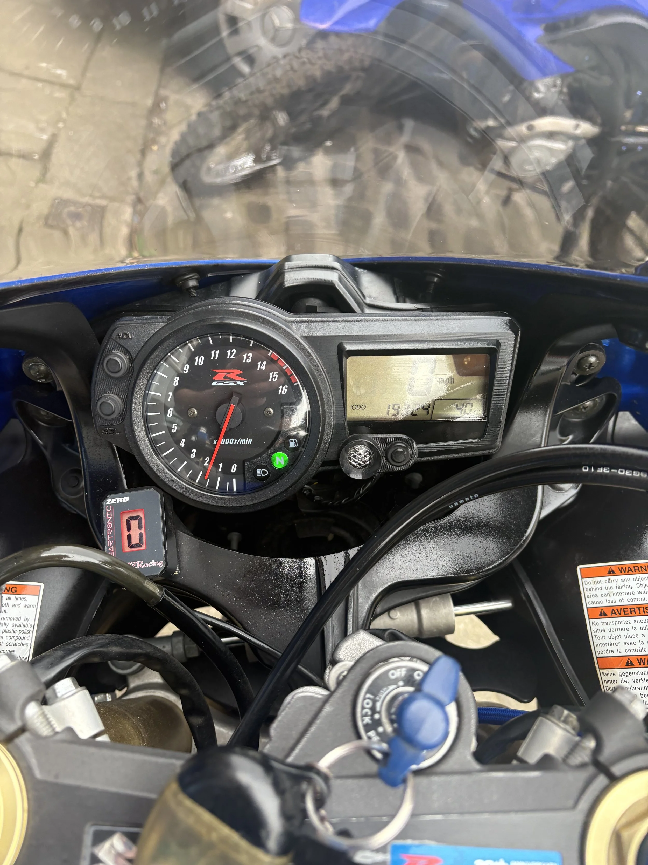 View from the rider's perspective on a blue motorcycle, showing the dashboard including a black and red tachometer, digital screen, ignition switch, and various warning labels.
