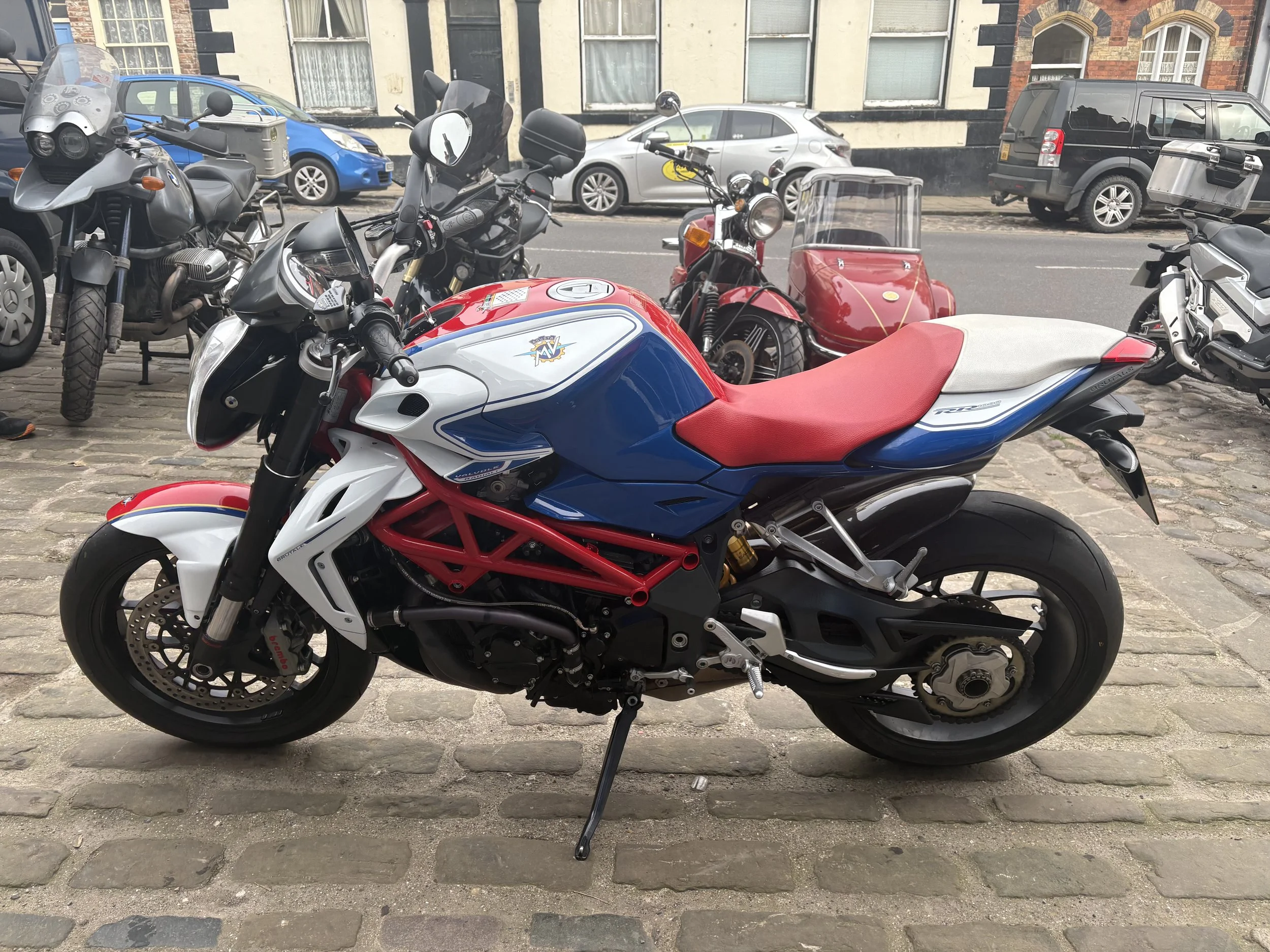 A Ducati motorcycle with a red, white, and blue color scheme parked on a cobblestone street, with other motorcycles and cars in the background.
