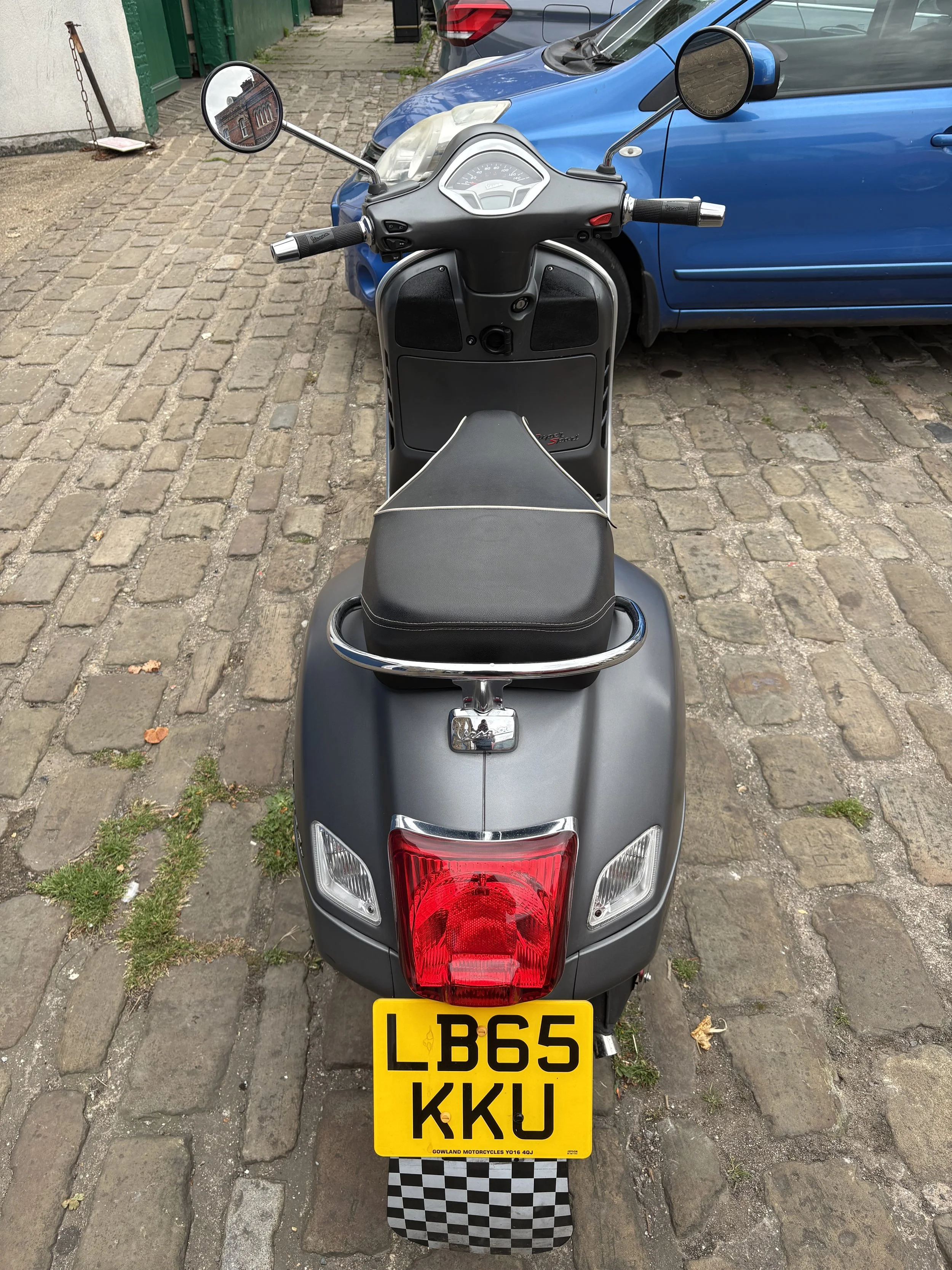 Rear view of a black scooter with a yellow license plate, parked on a cobblestone street next to a blue car.