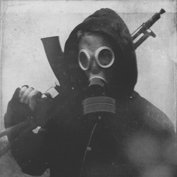 A person wearing a gas mask and a winter coat, holding a shovel with a rifle strapped to their back.