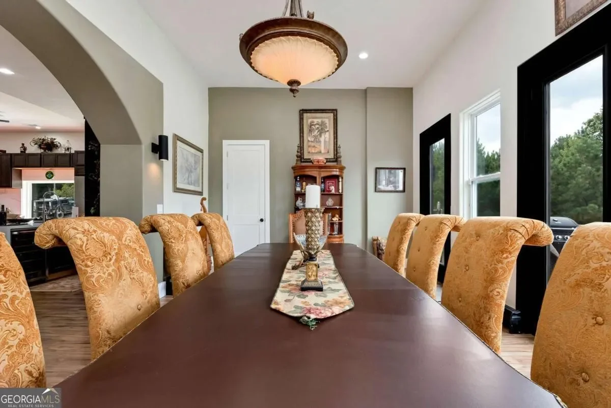 A dining room with a long wooden table, ornate yellow upholstered chairs, a floral table runner, and a chandelier. There are windows showing greenery outside and a wooden corner shelf with decorations.