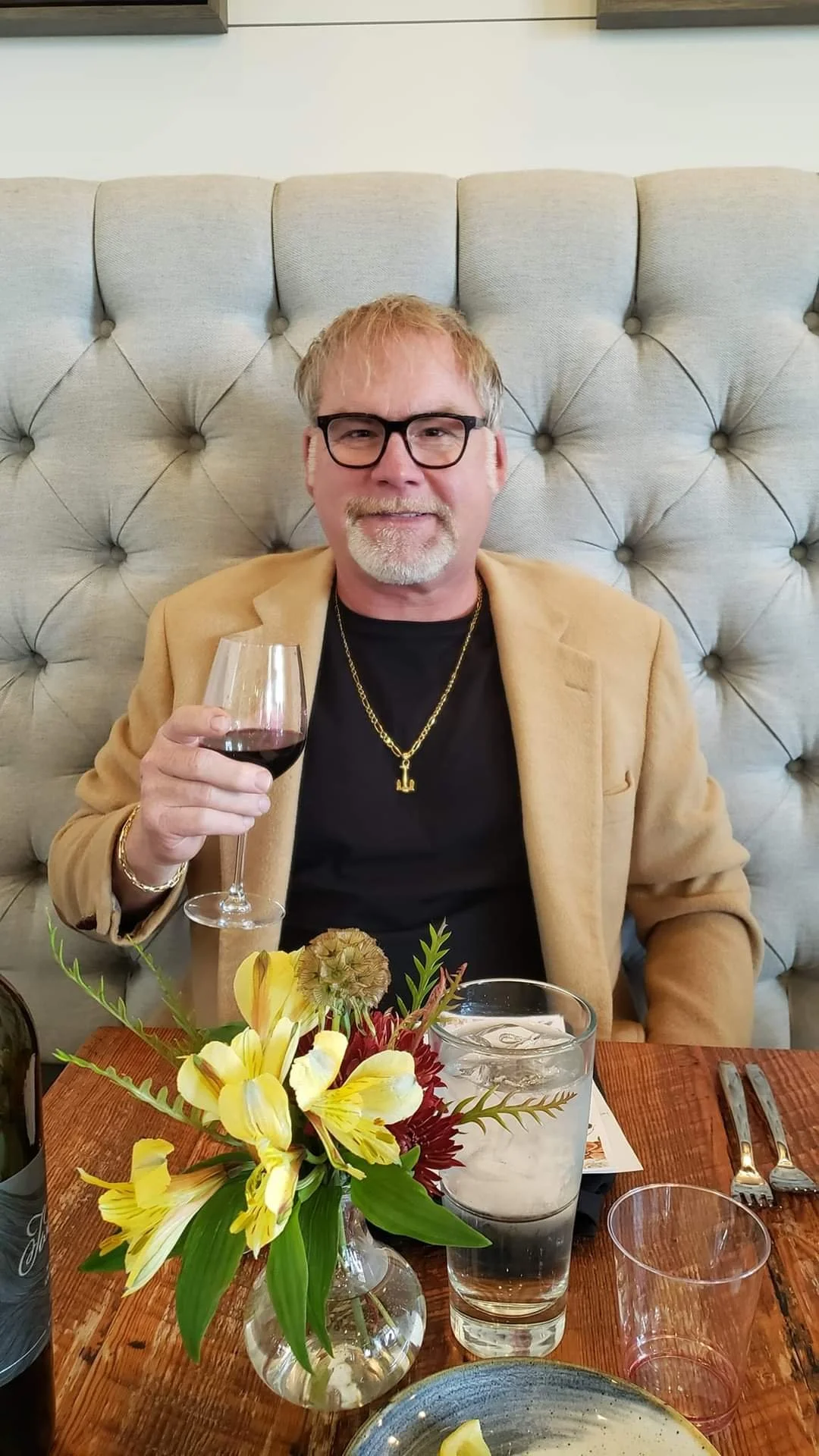 A middle-aged man with blond hair and glasses sitting at a restaurant table, holding a glass of red wine, wearing a black shirt, a tan blazer, and gold jewelry, with a flower arrangement, water glass, and utensils on the table.