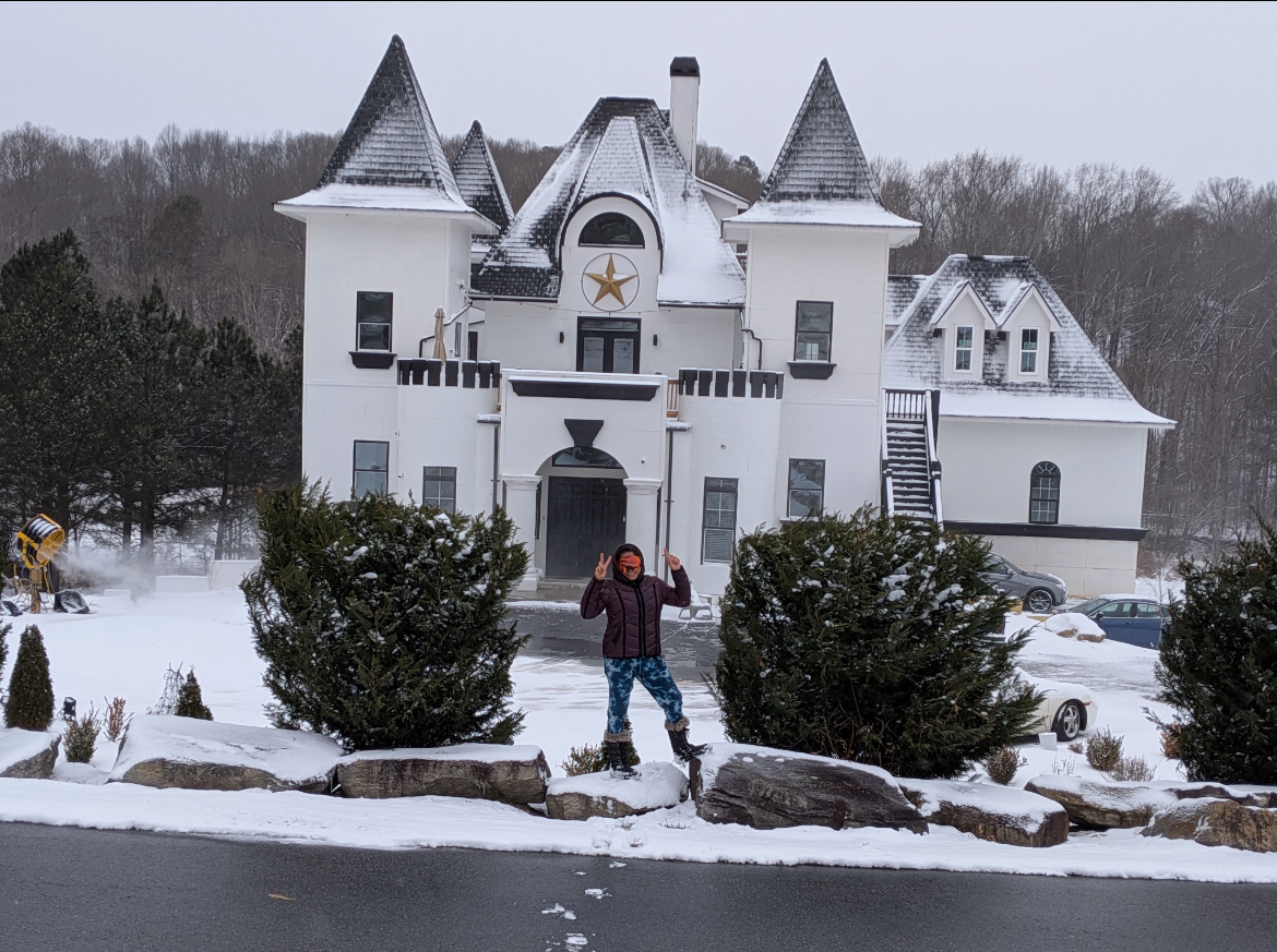 Person standing on snow-covered rocks in front of a large white castle-style house with pointed turrets and a star symbol above the entrance, surrounded by snow and trees.
