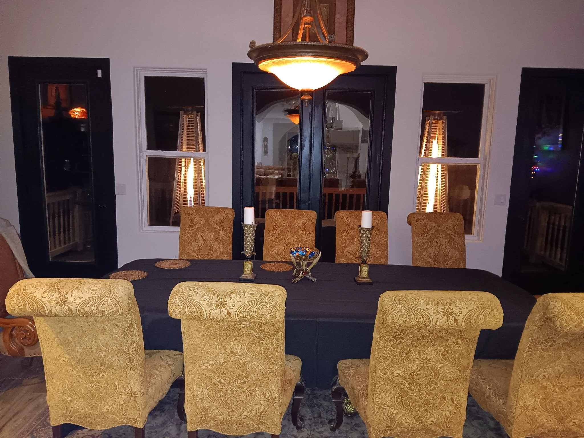 A dining room with a black covered table, eight gold-patterned chairs, a chandelier, and tall patio doors with blinds. Two large candles and a bowl of candies are on the table.