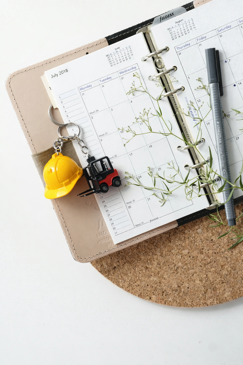 Agenda d'un calendrier avec un porte clé, un casque de chantier jaune, une petite grue rouge, un stylo noir, des branches de feuillage, et un sous-mug en liège.