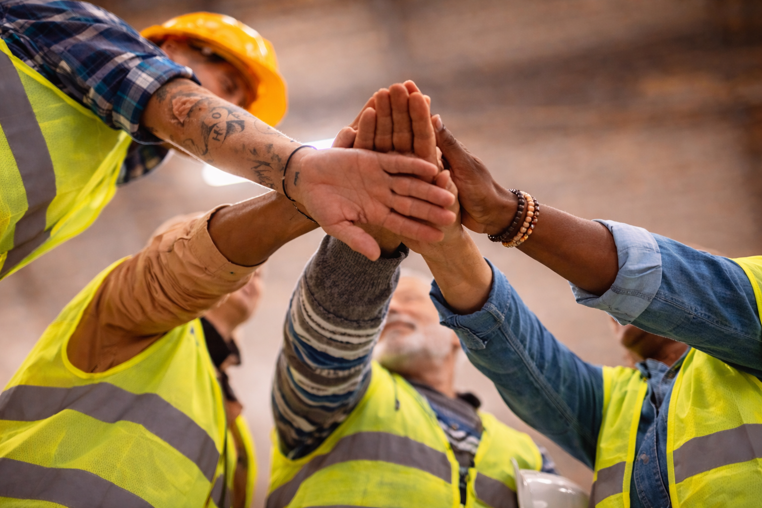 Groupe de travailleurs portant des gilets de sécurité jaune, faisant une accolade en se touchant les mains dans un espace intérieur.