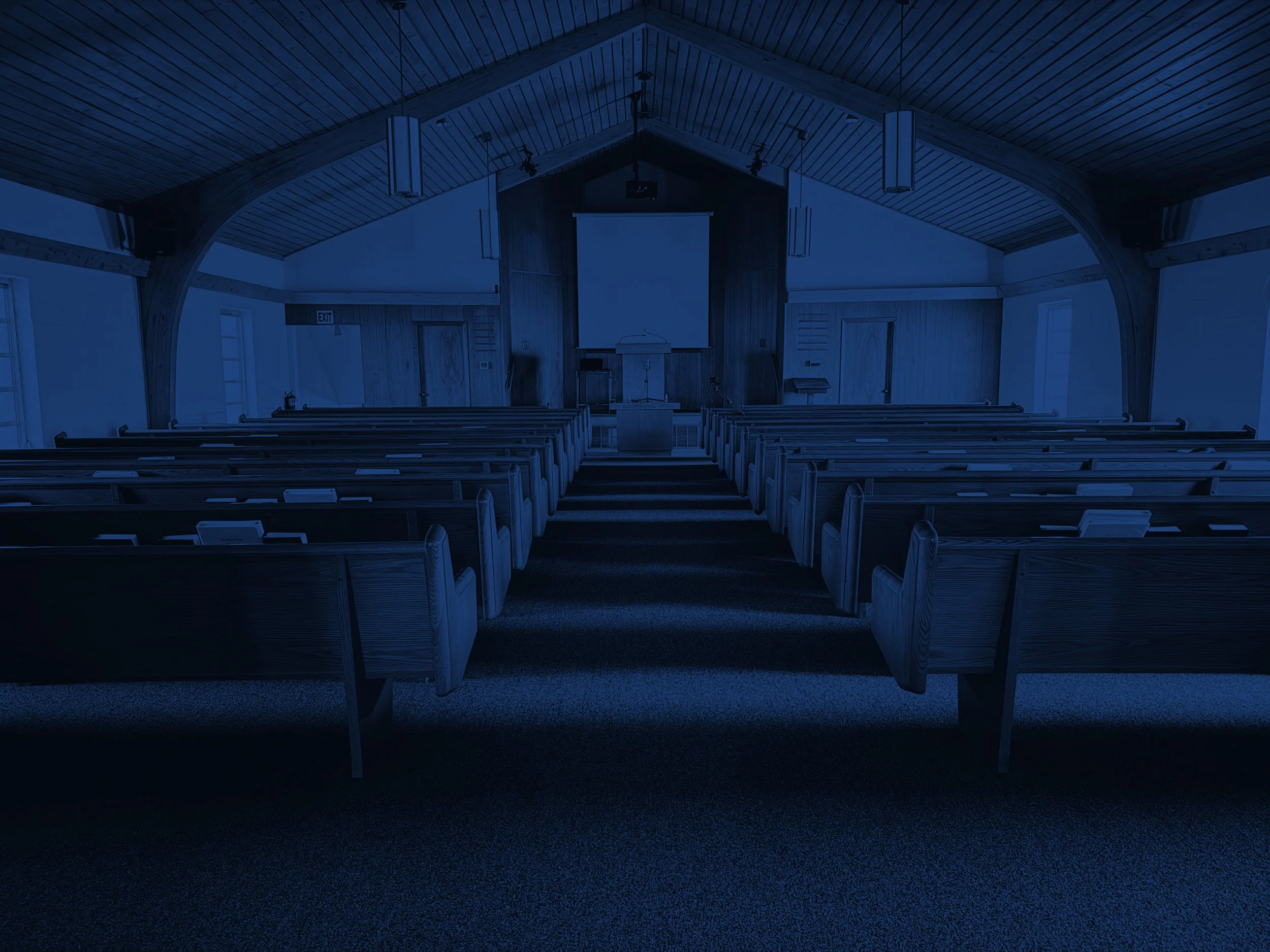 Interior of an empty church with wooden pews, a pulpit, and a large screen at the front, illuminated with dim blue lighting.