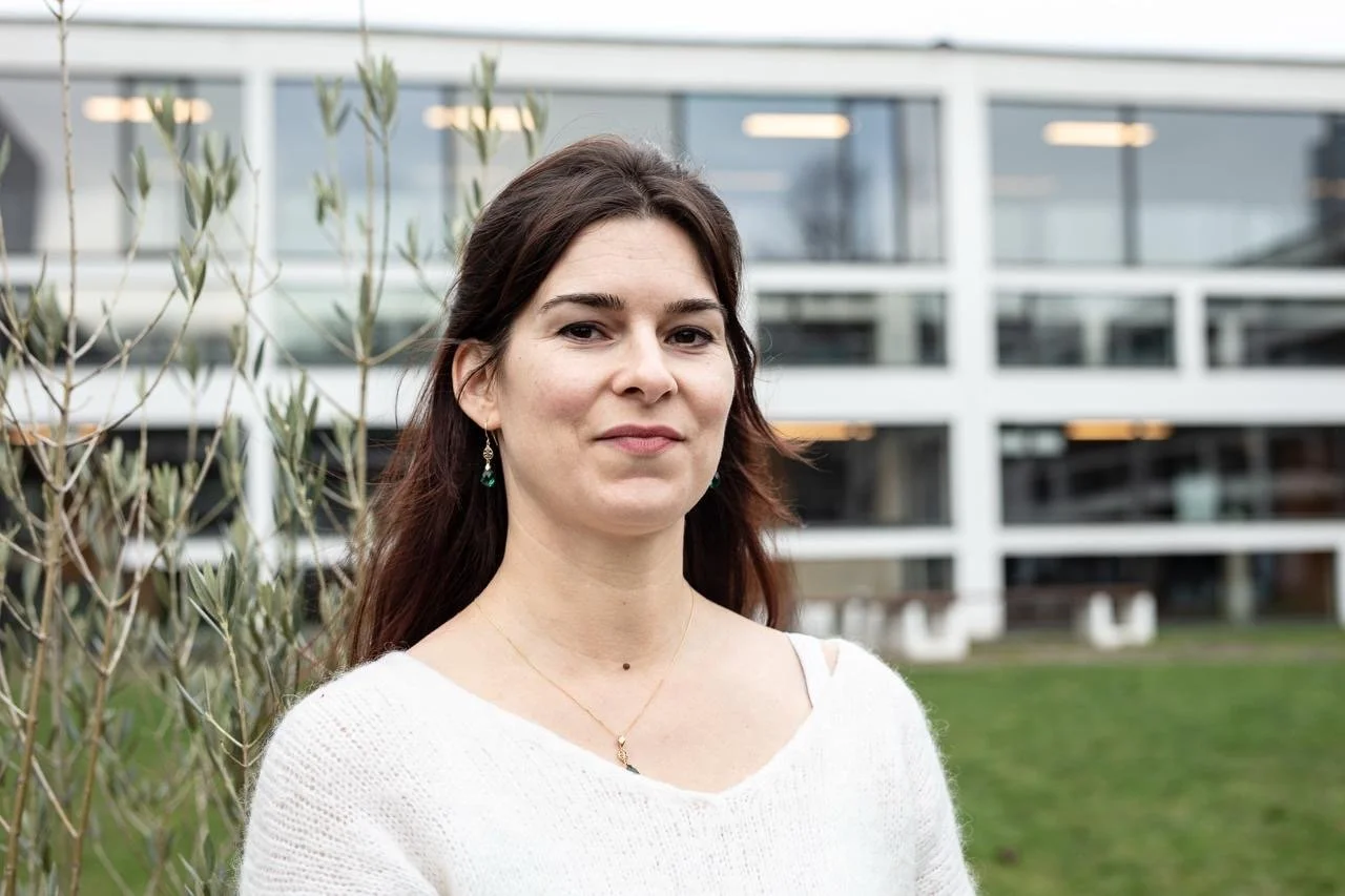 A woman with dark brown hair wearing a white sweater and earrings, standing outdoors with a modern glass building and greenery in the background.