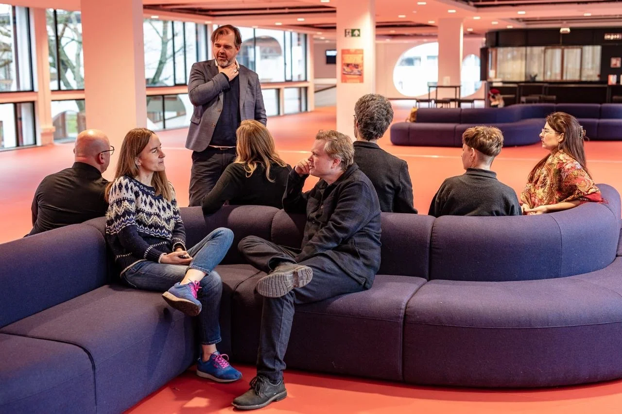A man standing and speaking to a group of seven people sitting in a curved purple sofa in a brightly lit, modern indoor space with large windows.