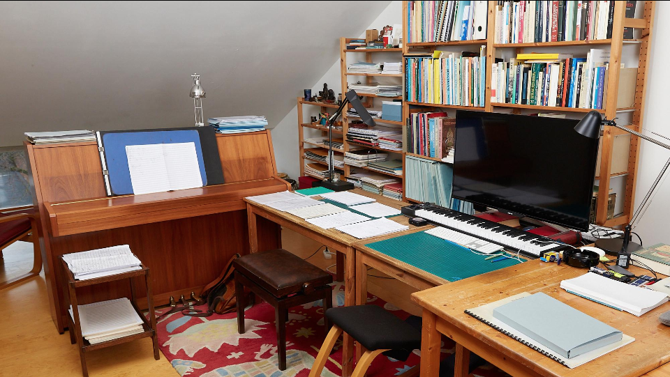A cozy home office with wooden furniture, a flat-screen TV, a keyboard, bookshelves filled with books and papers, a music stand, and a patterned rug.