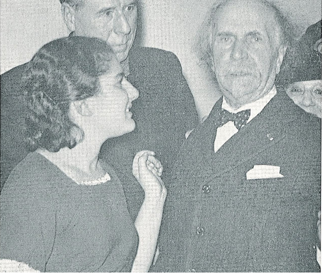 Four people in formal attire at a social gathering, smiling and interacting indoors.