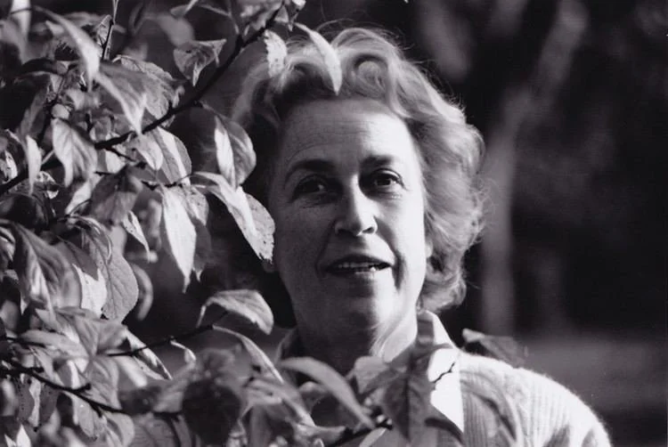 A black and white photo of a woman with light-colored, wavy hair, looking at the camera, with leaves partially covering her face, outdoors with trees in the background.