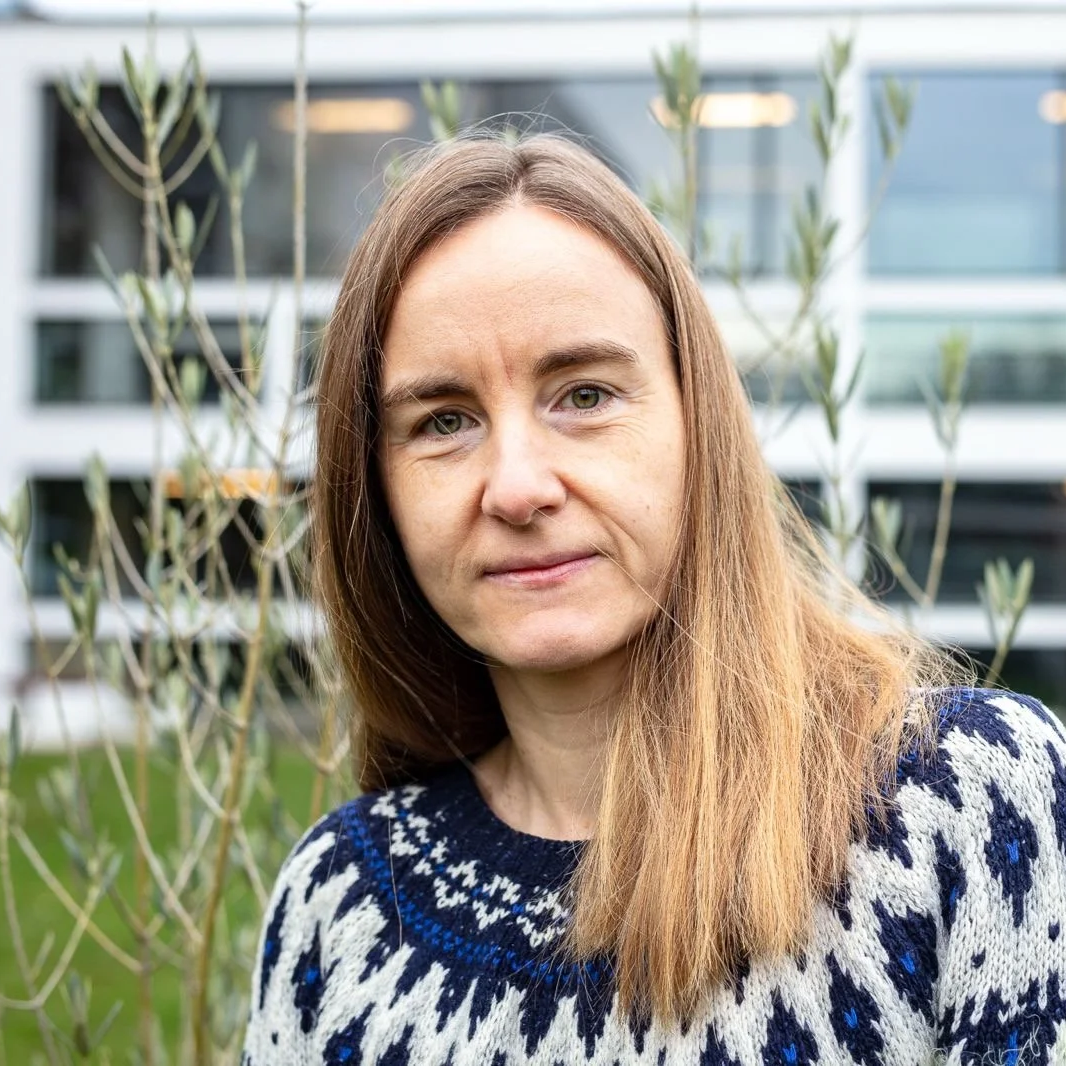 A woman with long light brown hair wearing a patterned sweater standing outdoors with a blurred building and green plants in the background.